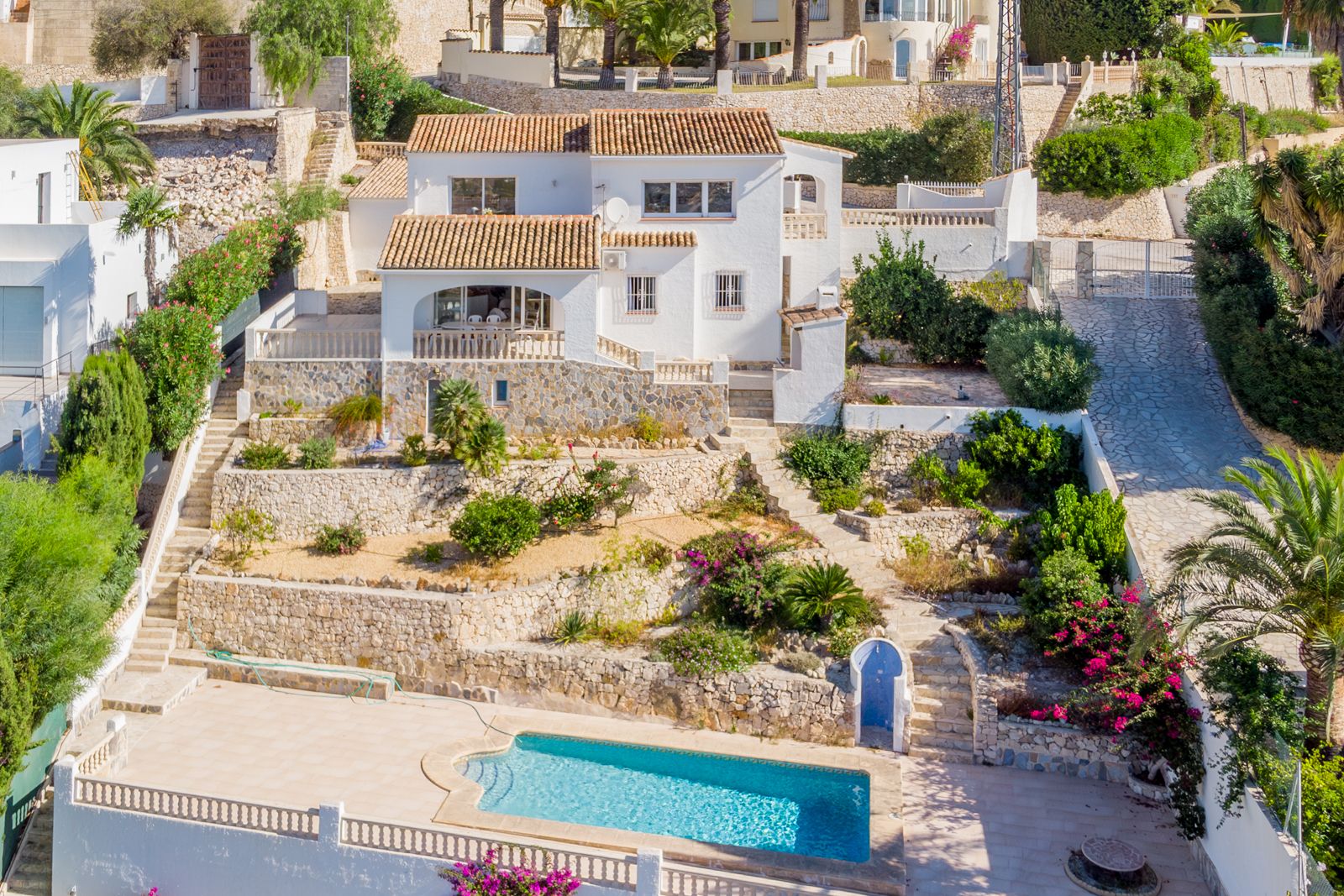 Vista exterior de la villa con fachada blanca, tejados de teja y jardín aterrazado con muros de piedra natural que conducen a la piscina privada.