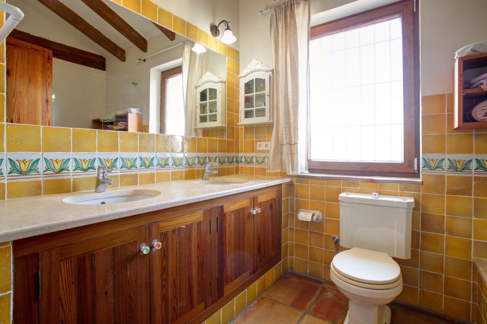 Baño rústico con encimera de mármol crema marfil, mueble de madera de mobila y azulejos mostaza. Incluye vigas de madera vistas y suelo de barro cocido tradicional.