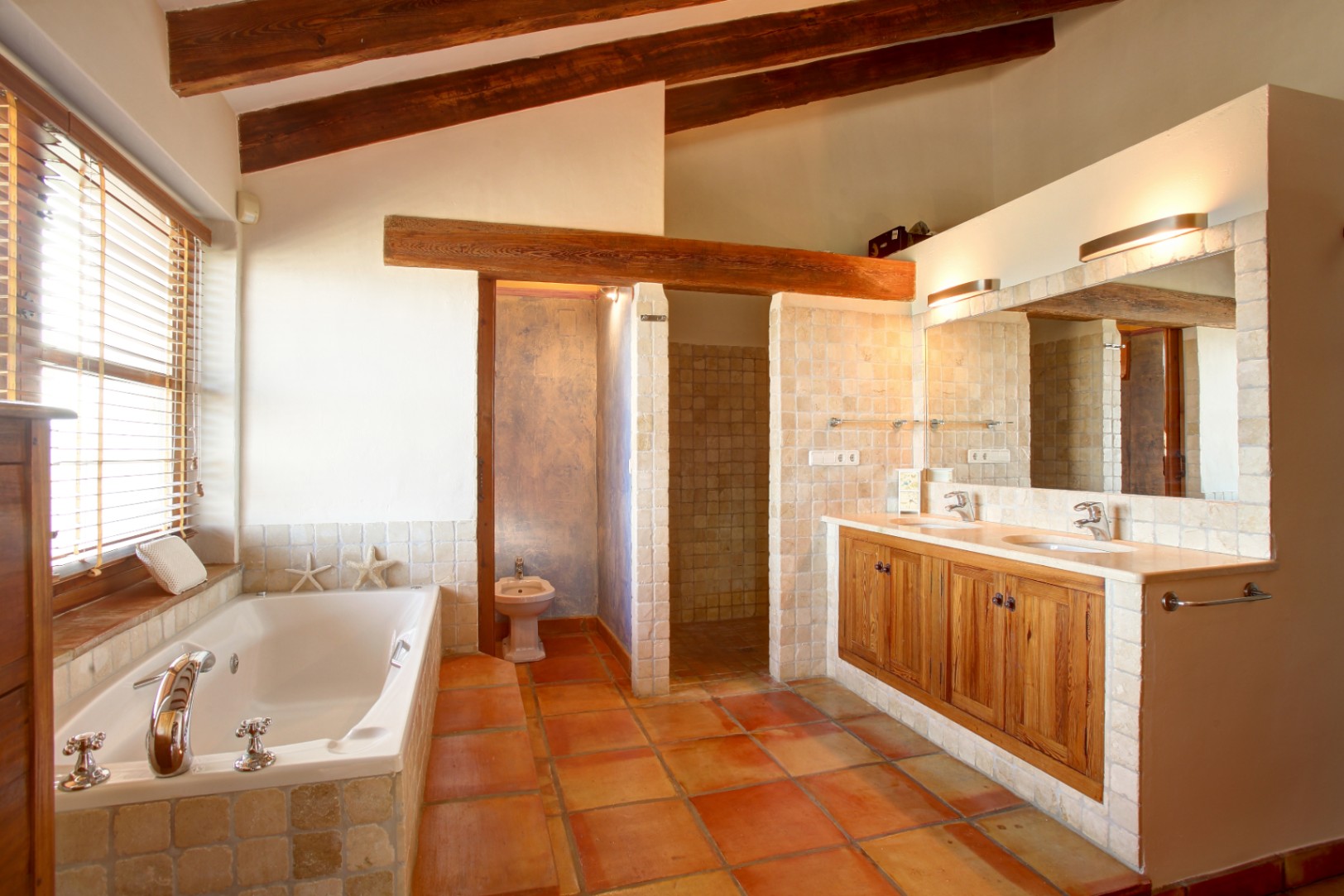 Baño rústico con vigas de madera vista, suelo de barro cocido y bañera encastrada. El mueble de lavabo doble combina madera de mobila con encimera de mármol y revestimiento de piedra natural.