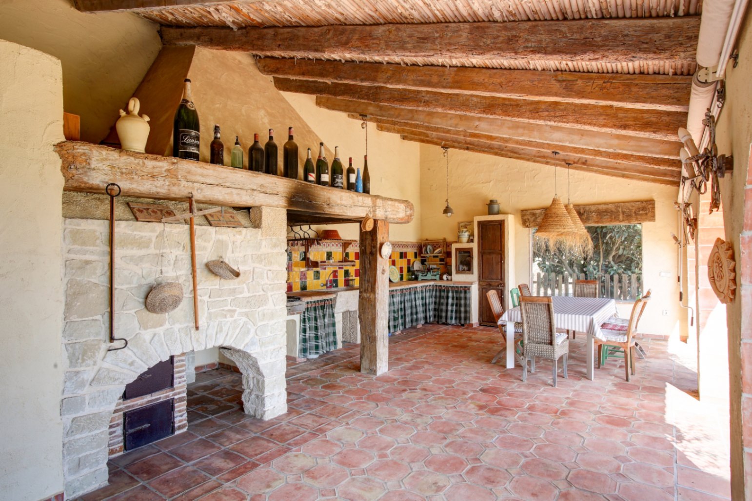Cocina de verano con barbacoa de piedra, vigas de madera recuperada y suelos de barro cocido. Incluye comedor exterior y azulejos tradicionales.