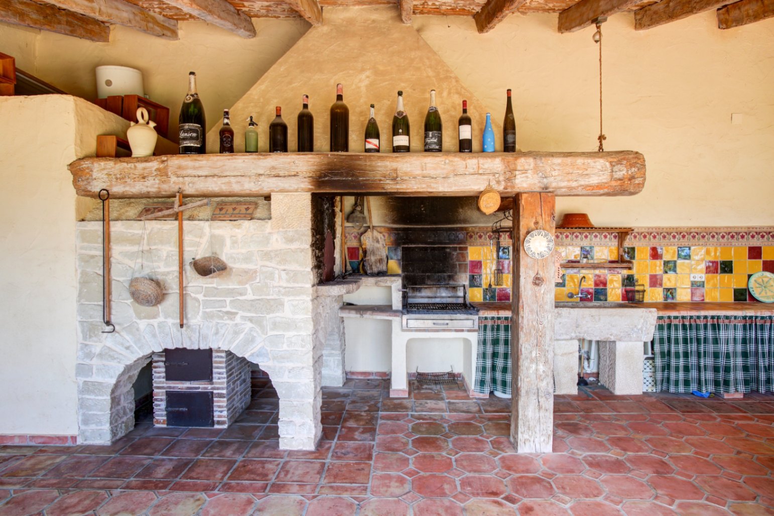 Cocina de verano rústica con barbacoa integrada, gran viga de madera recuperada, fregadero de piedra natural y azulejos artesanales sobre suelo de barro cocido.