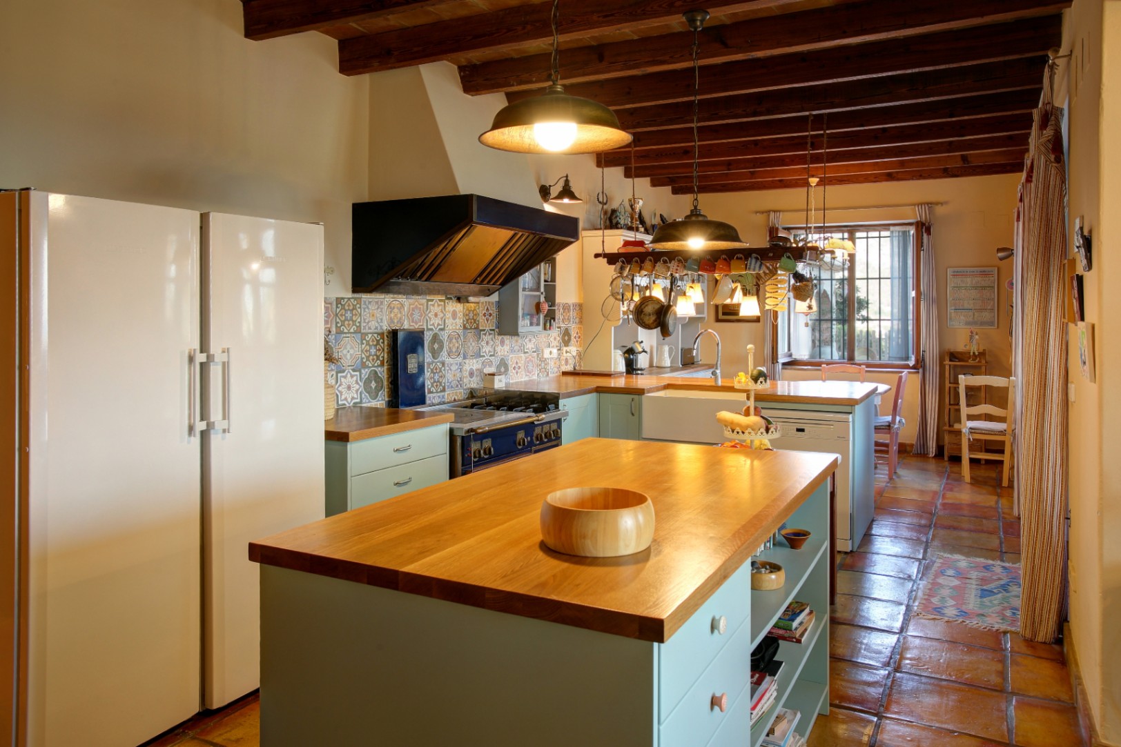 Cocina rústica con isla central de madera, vigas vistas y suelo de barro cocido. Destaca la cocina profesional azul, el salpicadero de azulejos decorativos y la abundante luz natural.