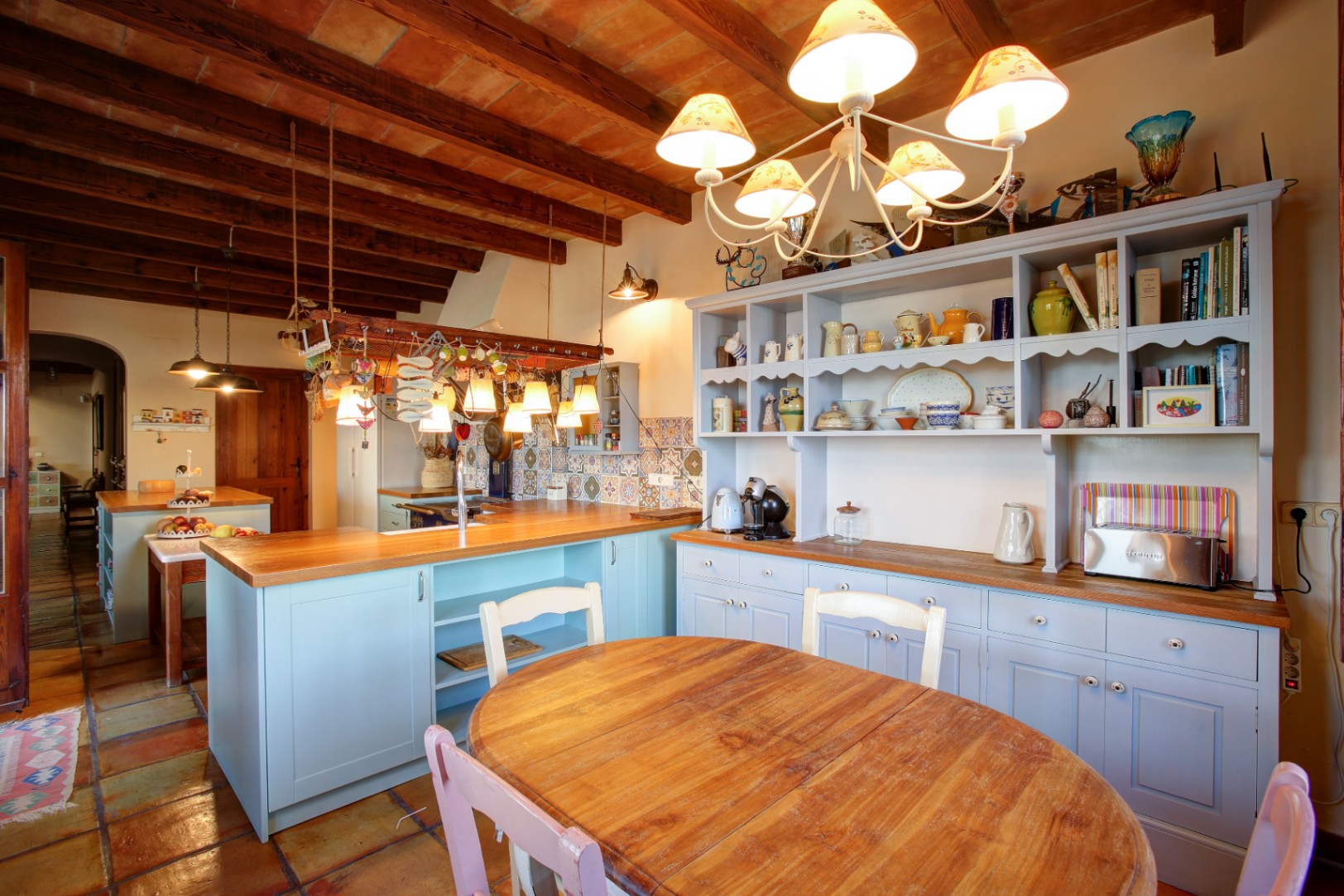 Cocina rústica con mobiliario azul claro, encimeras de madera y techos de vigas vistas con bovedillas. Incluye zona de comedor y suelos de terracota tradicional.