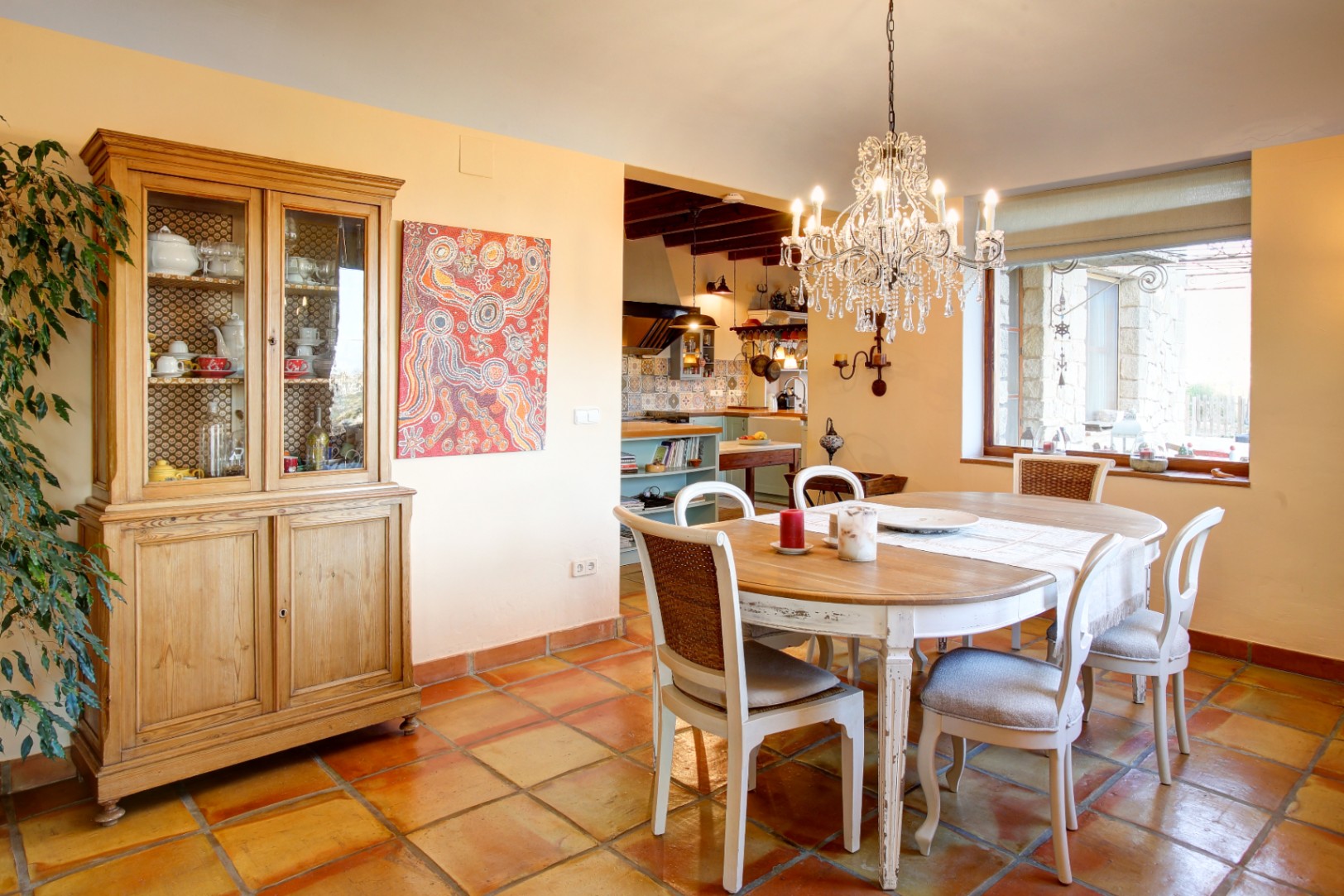 Comedor con mesa de madera y sillas decapadas bajo una lámpara de araña de cristal, con suelos de barro cocido y alacena de madera natural.