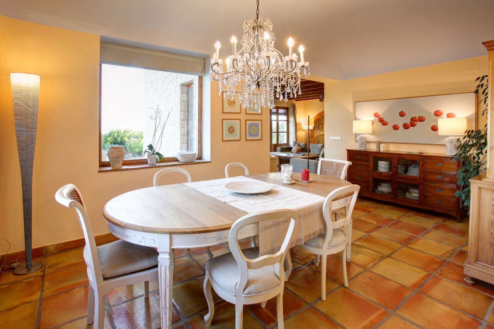 Comedor rústico con suelo de barro cocido, mesa de madera decapada y lámpara de araña de cristal. El ventanal ilumina el espacio y muestra los muros de piedra exteriores.