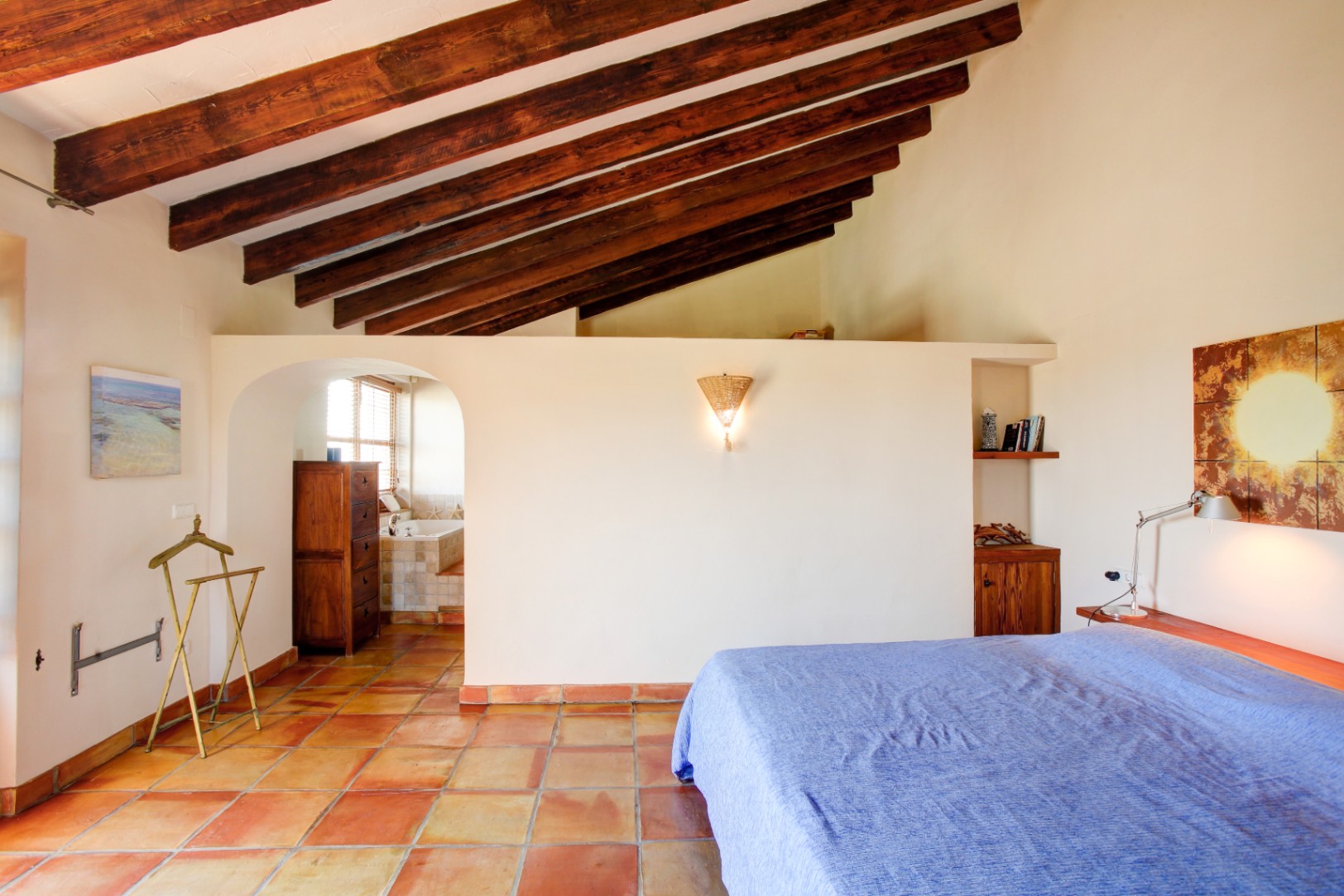Dormitorio principal con techos altos de vigas de madera vista, suelos de barro cocido y acceso directo a un baño en suite a través de un arco arquitectónico.