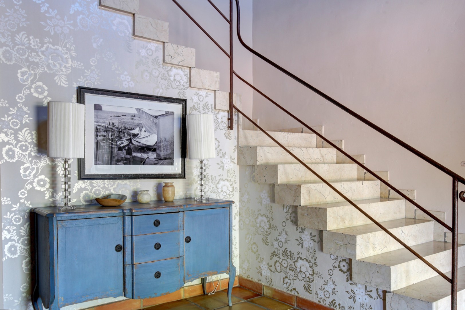 Escalera de mármol macizo con barandilla metálica, junto a un aparador azul decapado sobre suelo de barro cocido y pared con papel pintado floral.