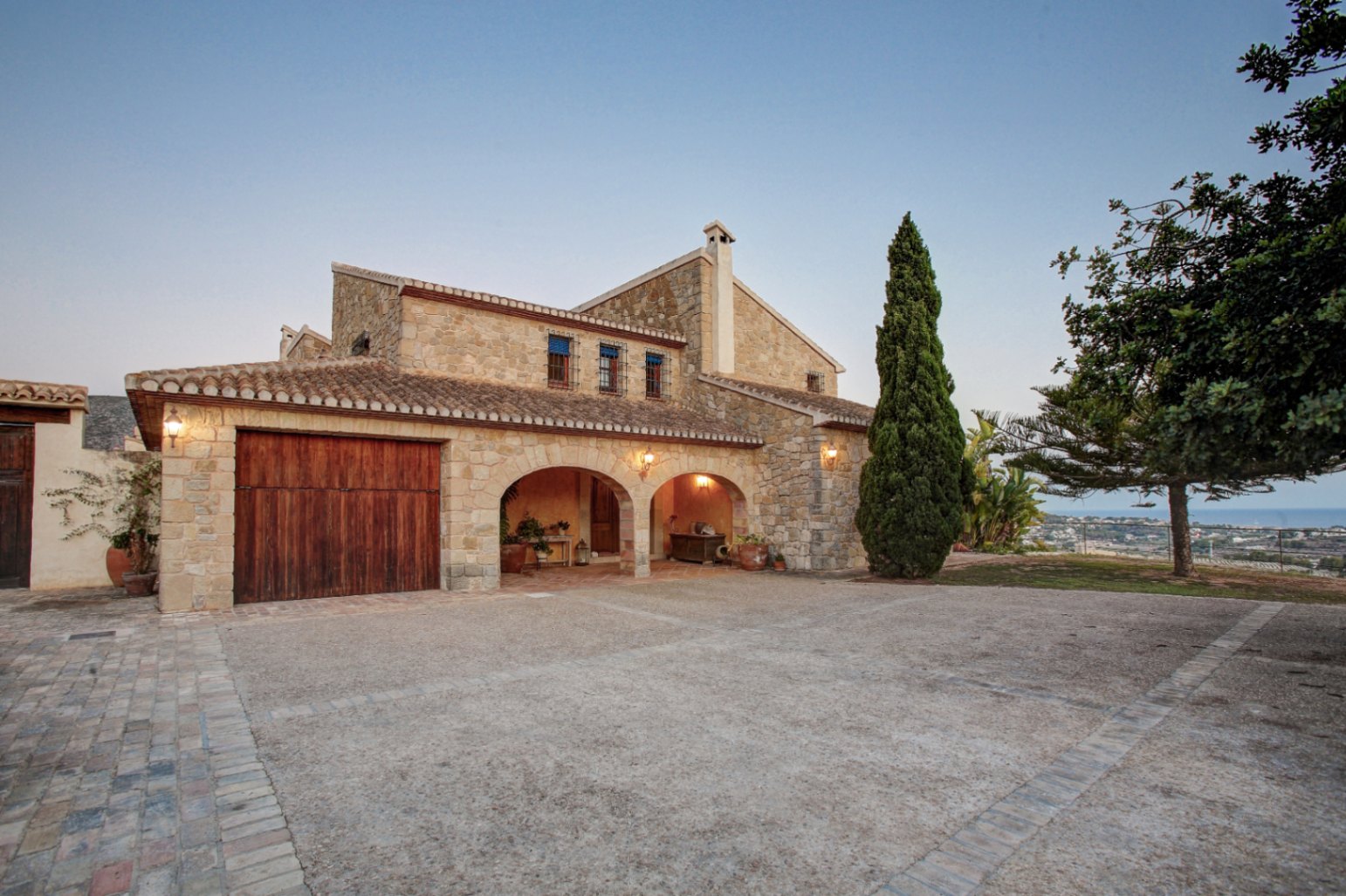 Fachada de piedra natural con arcos rústicos, garaje de madera maciza y amplio patio pavimentado con vistas despejadas al mar Mediterráneo.