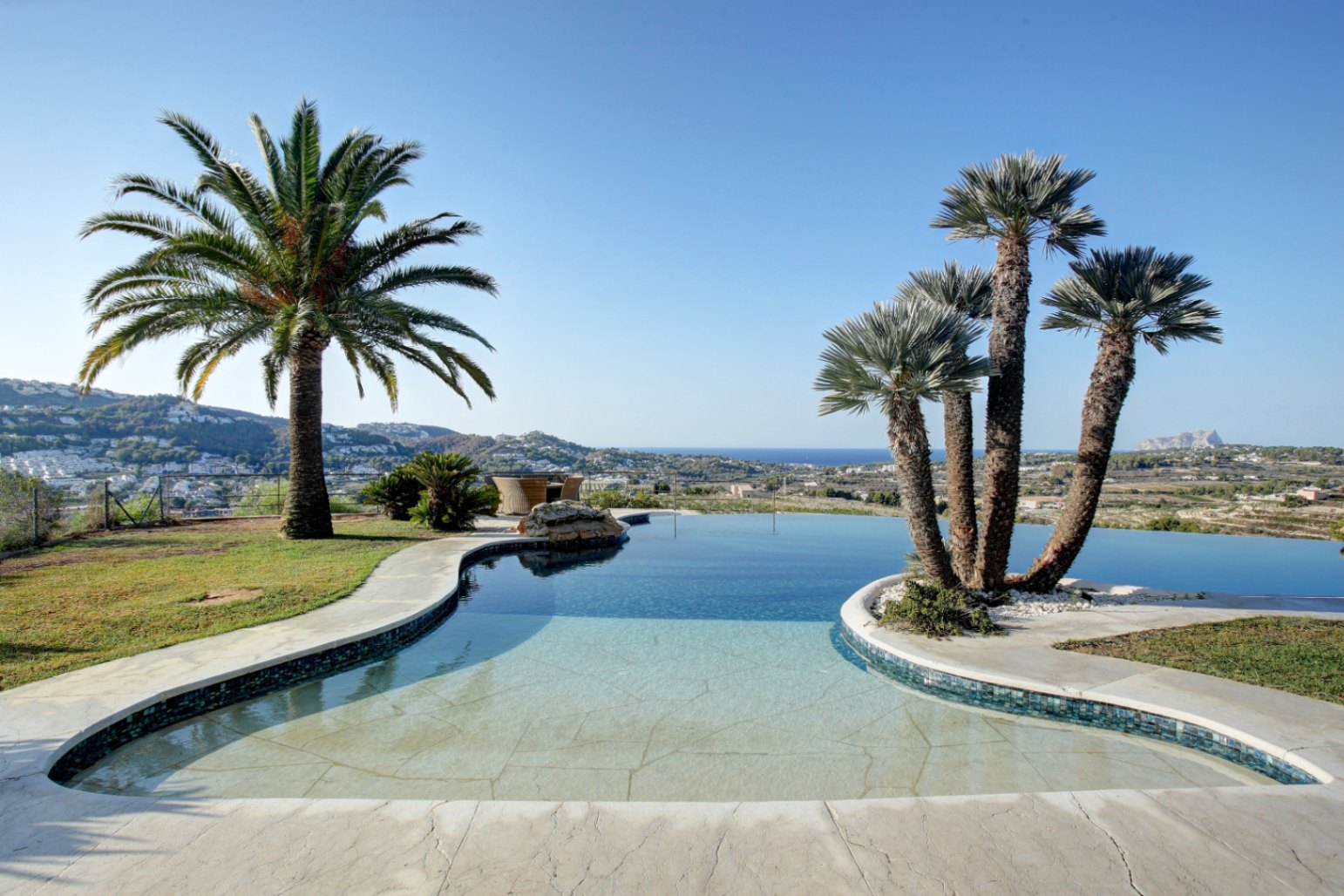 Piscina de diseño orgánico con borde infinito, rodeada de palmeras y vegetación mediterránea, con vistas panorámicas al mar y a la costa de Moraira bajo un cielo despejado.