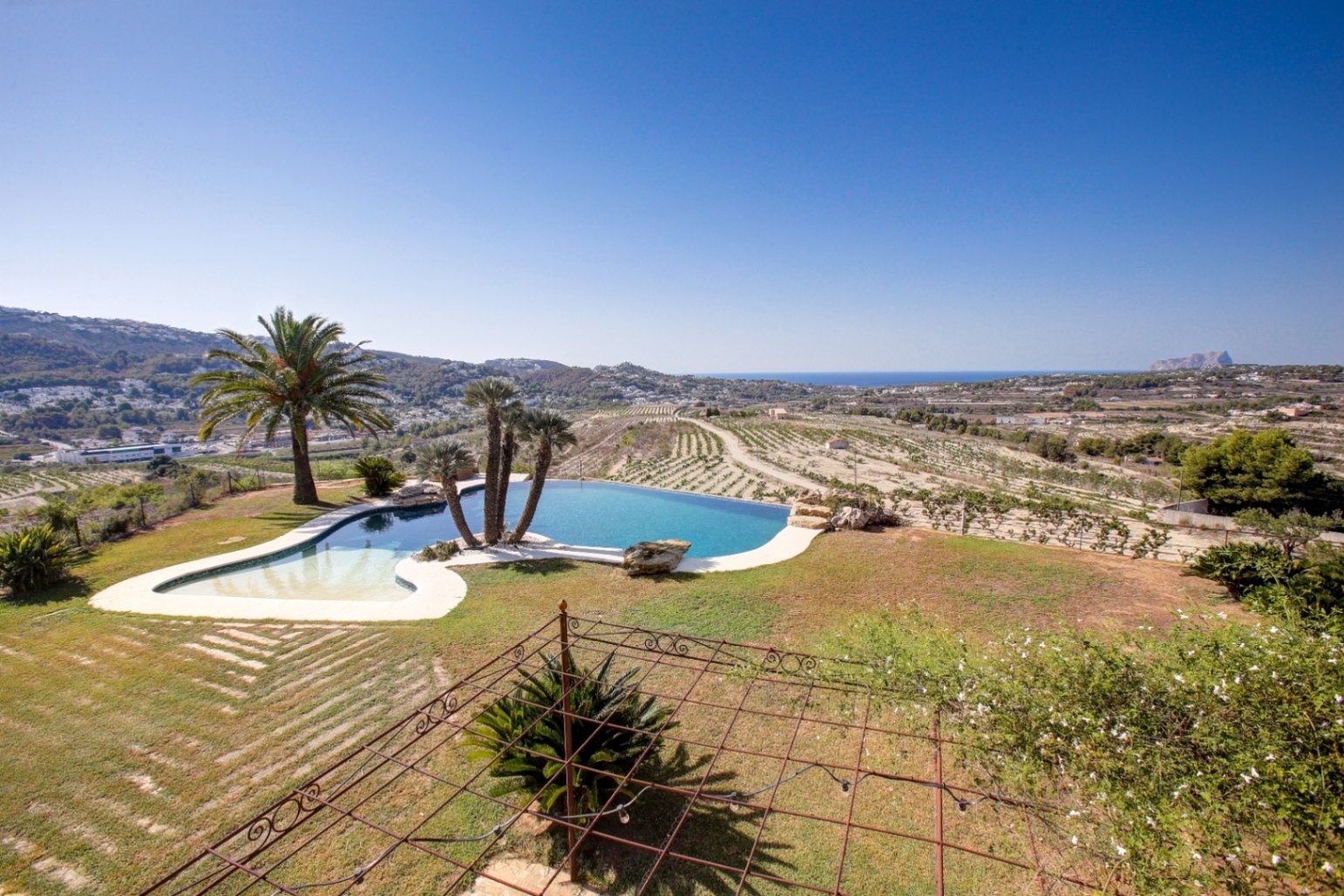 Piscina de diseño orgánico con vistas panorámicas al mar y al Peñón de Ifach, rodeada de amplias zonas verdes y terrenos de cultivo mediterráneo.