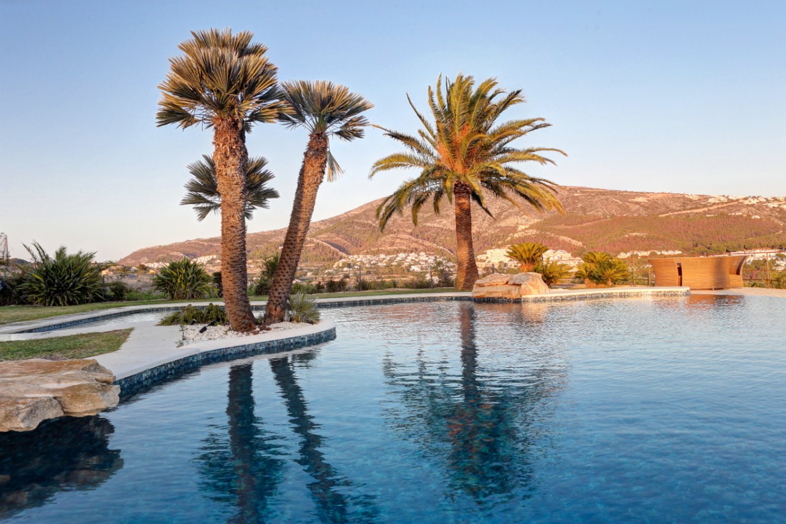 Piscina de forma orgánica con gresite azul oscuro, rodeada de palmeras y rocas naturales, con vistas panorámicas a las montañas y al valle bajo la luz del atardecer.