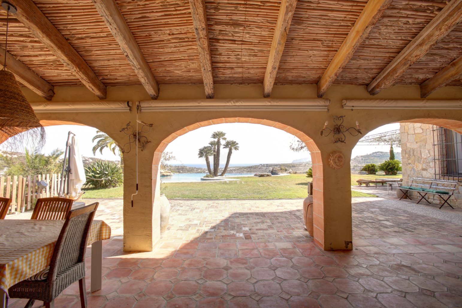 Porche cubierto con vigas de madera, techo de cañizo y suelos de barro cocido. Los arcos enmarcan vistas al jardín, la piscina con palmeras y el mar Mediterráneo.