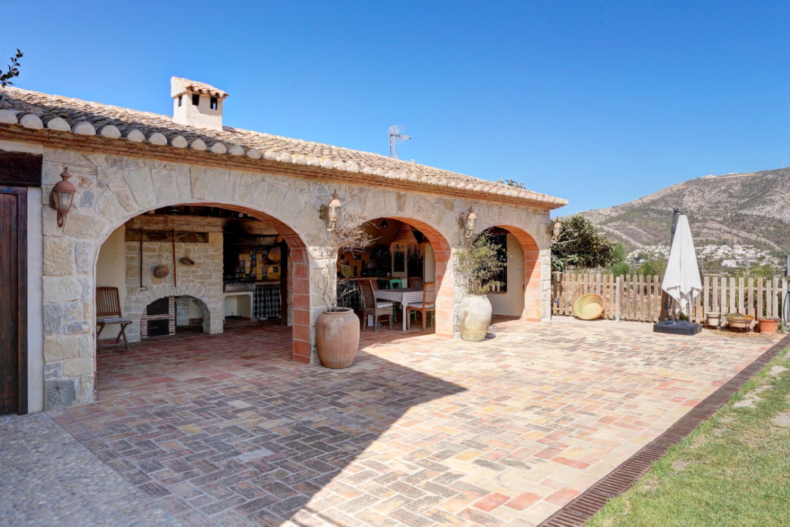Porche rústico con arcos de piedra y ladrillo, suelo de barro cocido y zona de barbacoa tradicional integrada frente a un paisaje de montaña.
