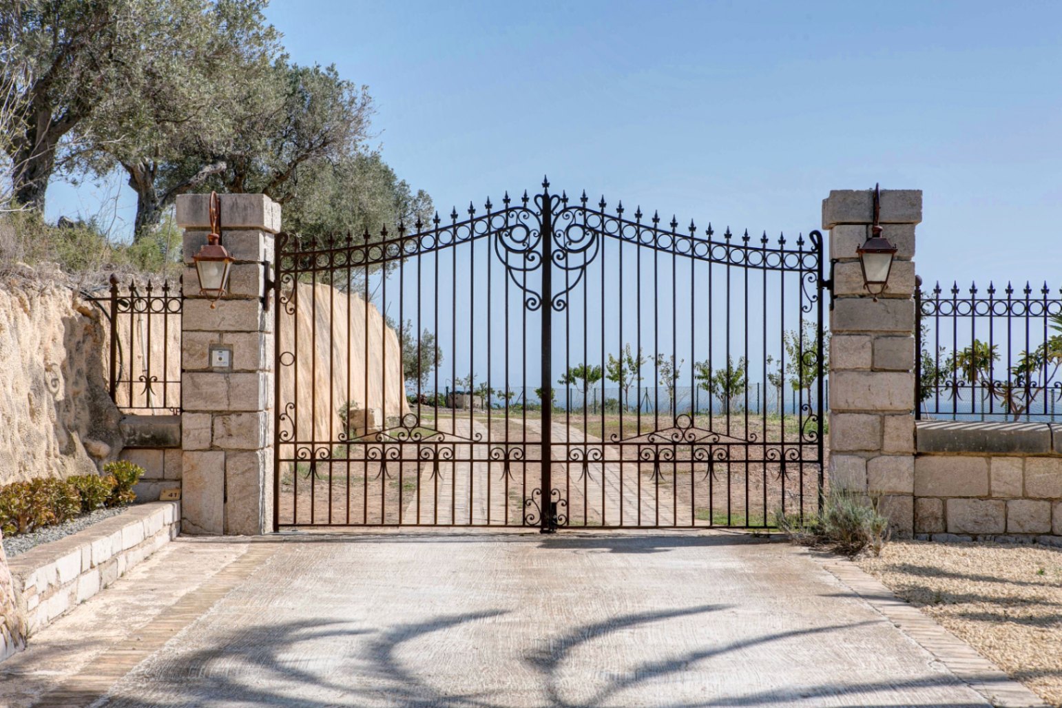 Puerta de entrada principal de hierro forjado ornamentado, flanqueada por pilares de piedra natural con faroles de estilo clásico y acceso pavimentado.