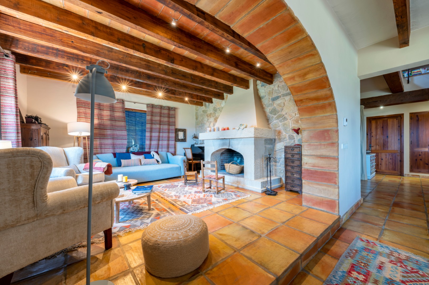Salón rústico con chimenea de piedra, techos de vigas de madera y bovedillas, suelos de barro cocido y un gran arco divisorio de ladrillo visto.