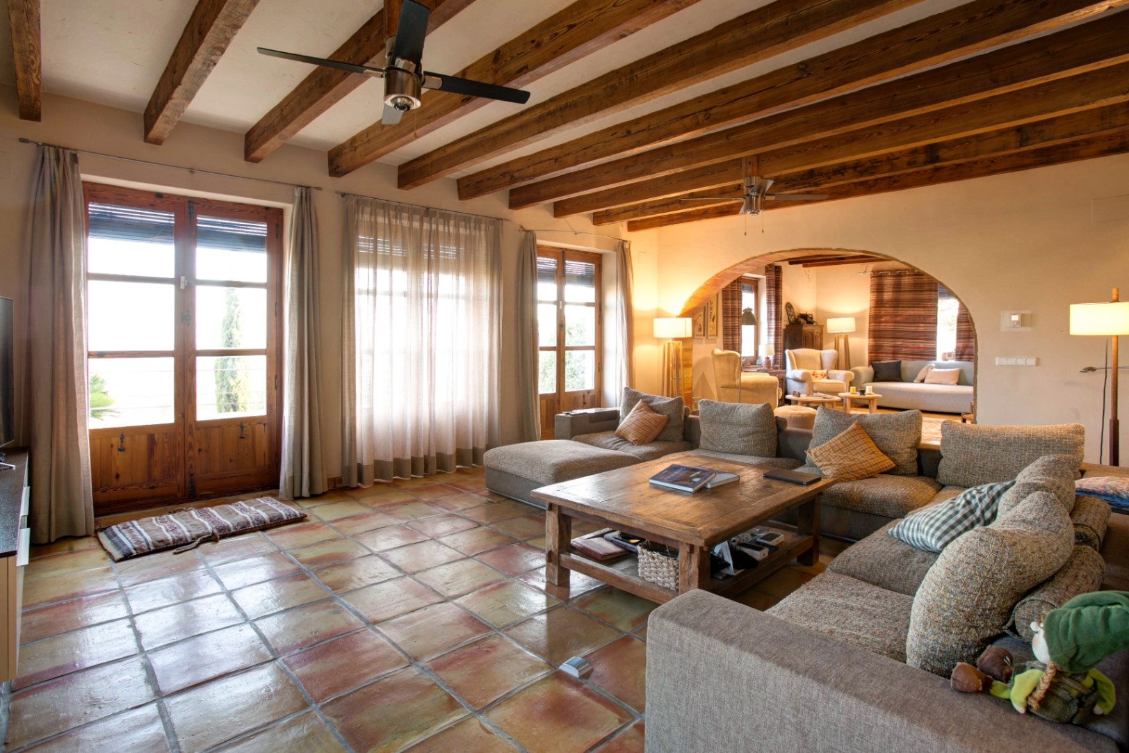 Salón rústico con suelos de barro cocido, vigas de madera vista y un arco que comunica ambientes. Amplios ventanales de madera inundan de luz natural la zona de estar.