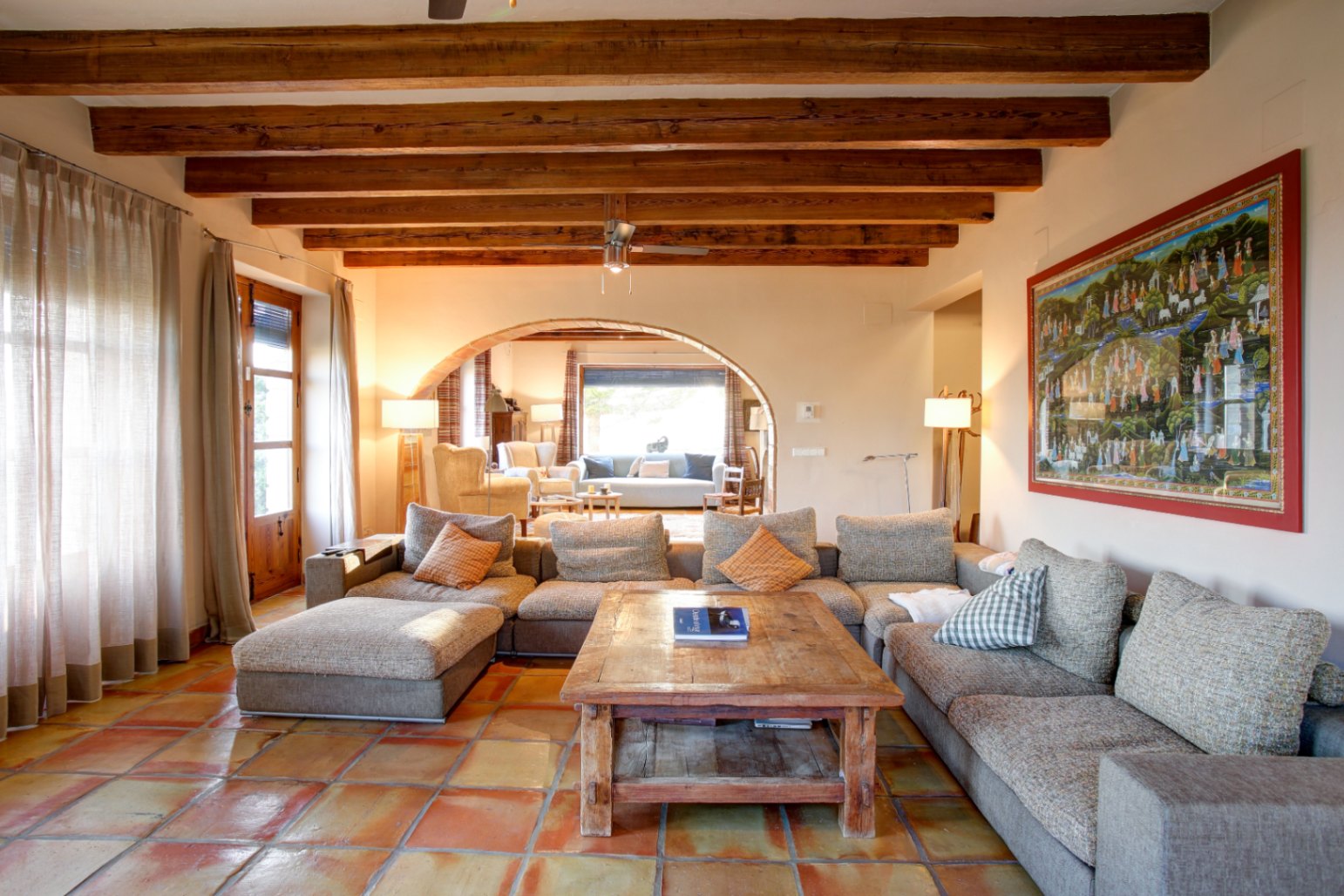 Salón rústico con vigas de madera vista, suelos de barro cocido y un gran sofá modular. Un arco de medio punto comunica con una segunda zona de estar luminosa.