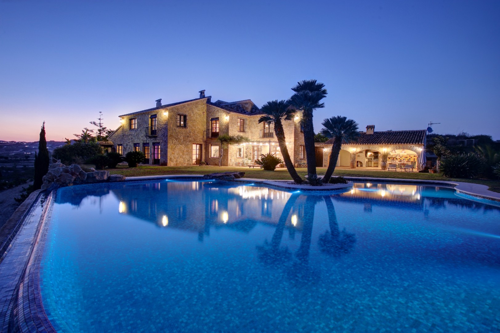 Vista nocturna de la fachada de piedra y la piscina iluminada, con la cocina de verano al fondo y palmeras integradas en el entorno paisajístico.