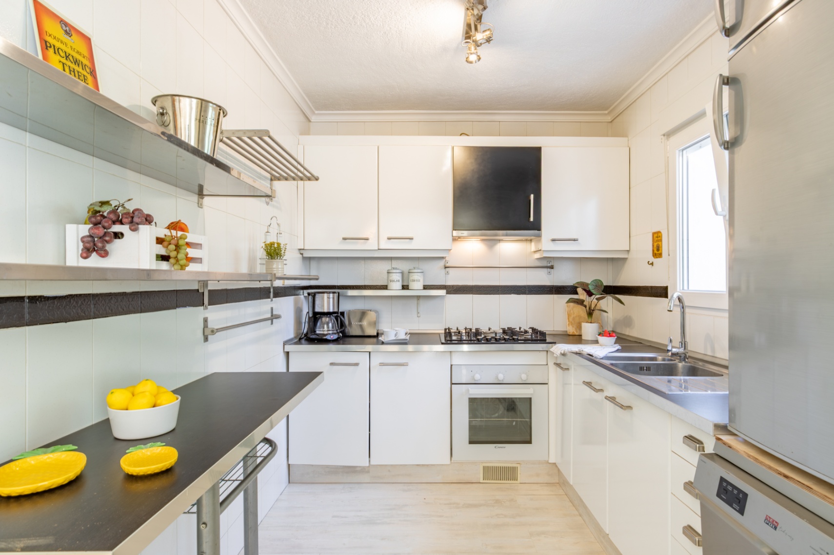 Cocina equipada con mobiliario blanco, encimeras oscuras, estanterías de acero inoxidable y luz natural. Incluye placa de gas, horno y fregadero doble de acero.