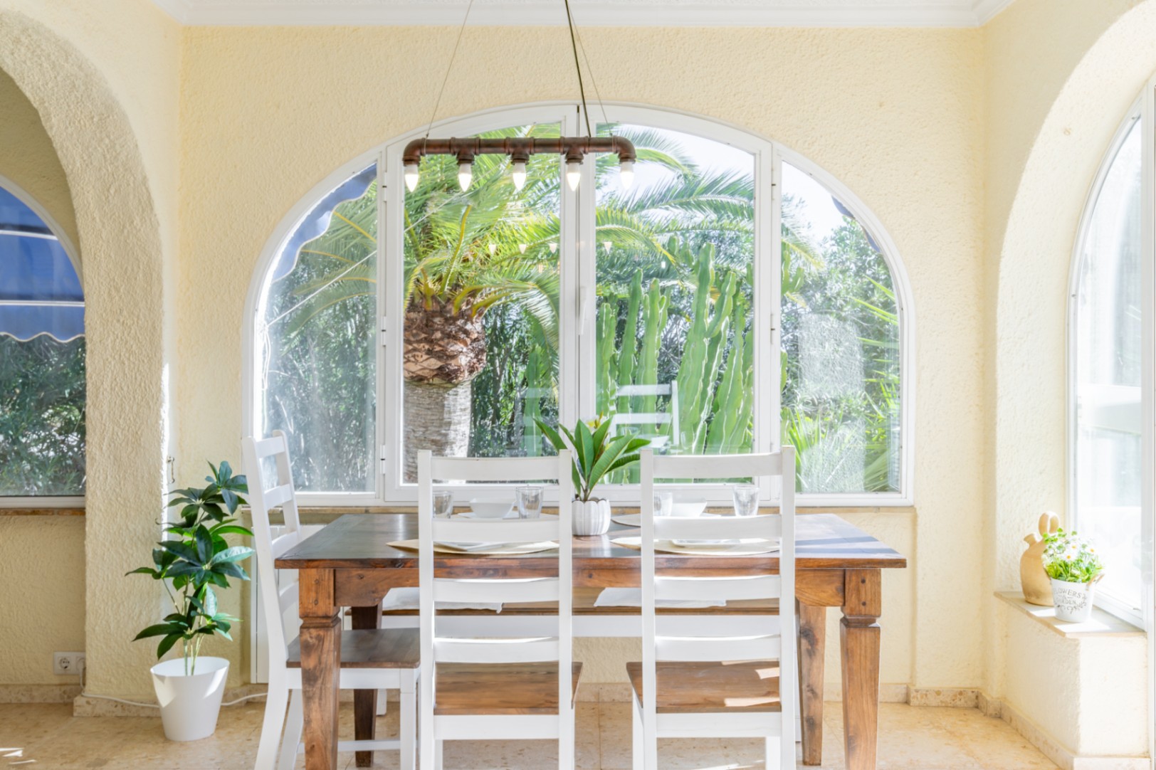 Comedor luminoso en terraza acristalada con mesa de madera, sillas blancas y lámpara industrial, ofreciendo vistas al jardín tropical con palmeras.