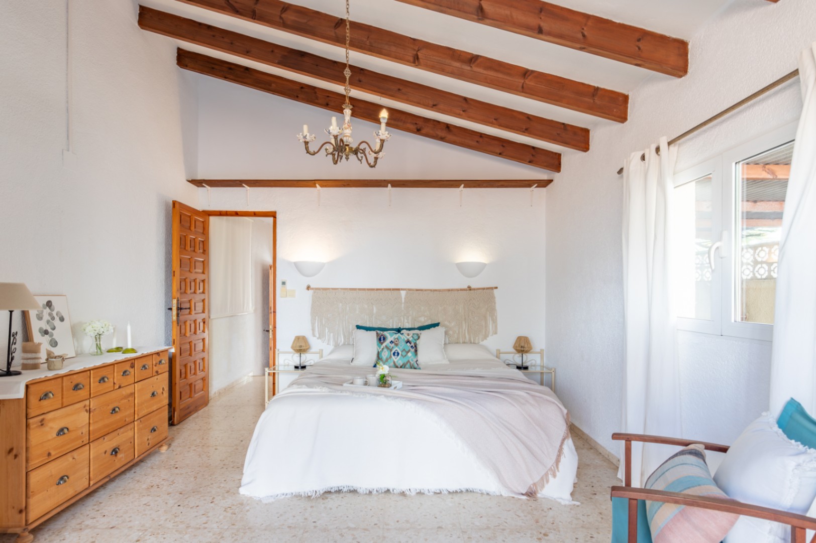 Dormitorio con techos altos de vigas de madera expuestas, suelos de terrazo y una decoración mediterránea con detalles en macramé y mobiliario de madera natural.