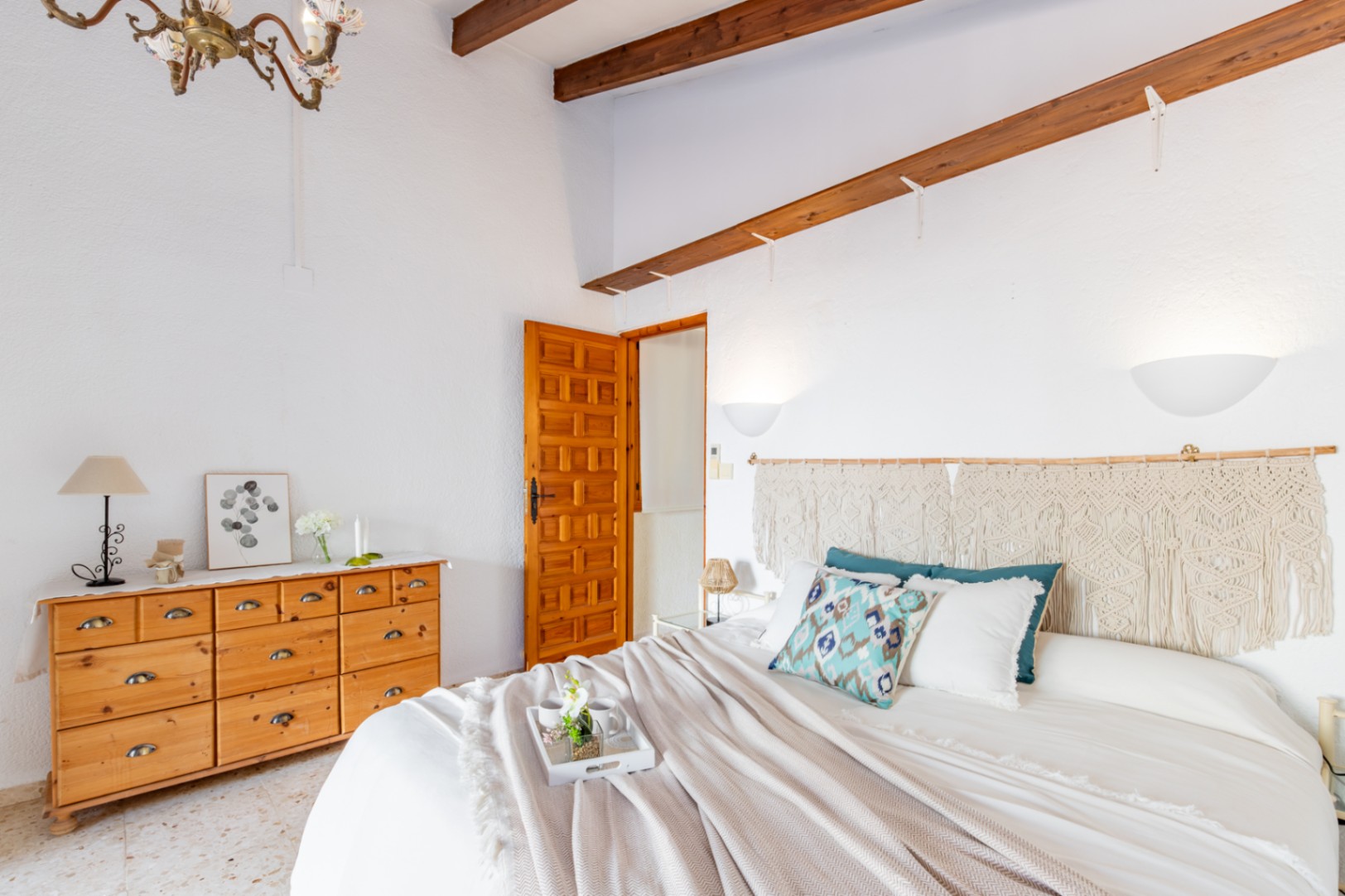 Dormitorio con vigas de madera vistas, cómoda de pino y cabecero de macramé. Incluye suelos de terrazo original y acceso a través de una puerta de madera tallada de estilo rústico.