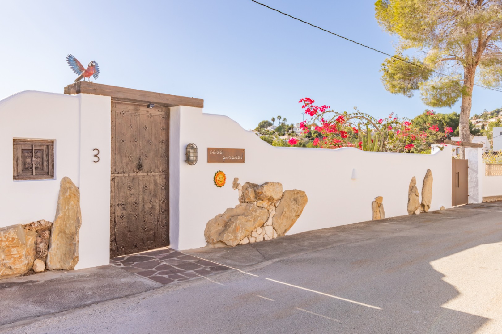 Entrada principal con puerta de madera rústica y herrajes, integrada en un muro blanco con detalles de piedra natural y buganvillas bajo un cielo despejado.