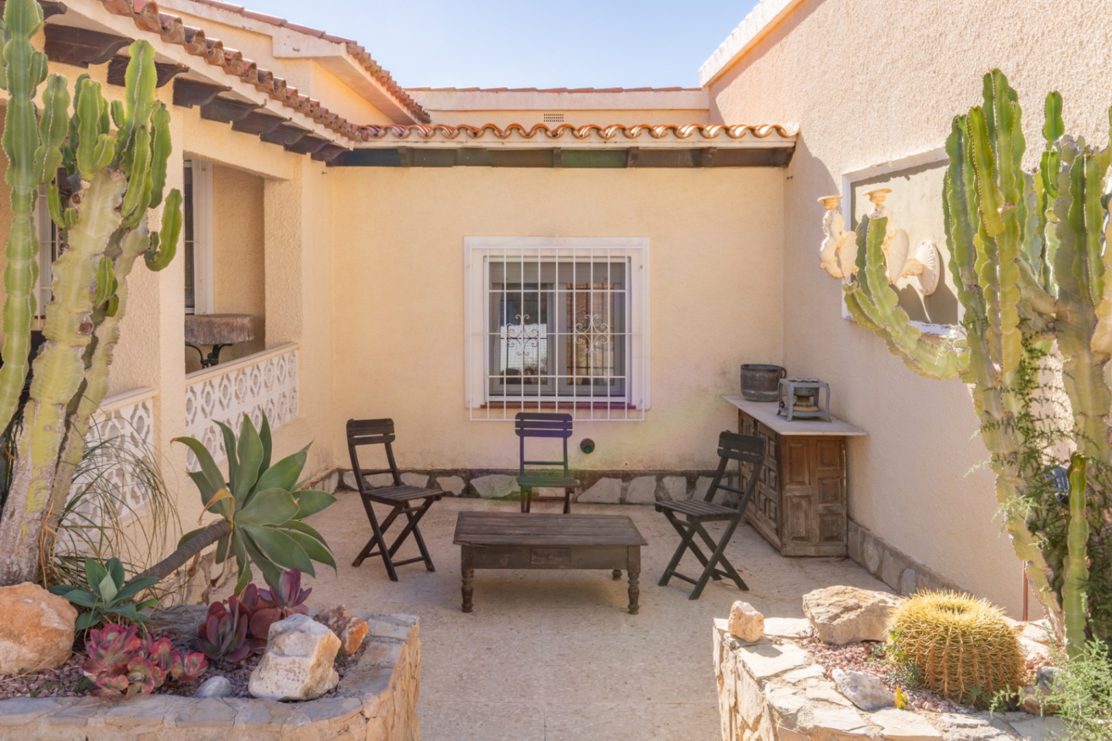 Patio exterior de estilo mediterráneo con suelo de piedra, mobiliario de madera rústica y jardineras integradas con cactus y suculentas bajo el sol de Moraira.