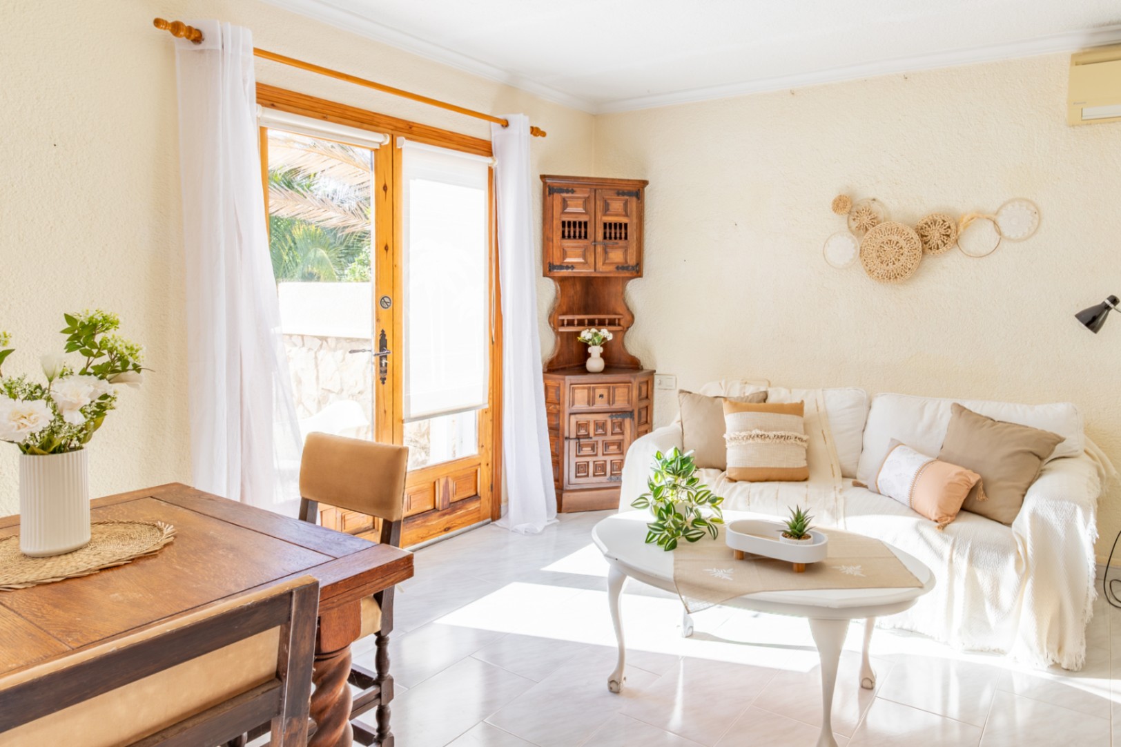 Salón comedor luminoso con mobiliario rústico de madera, sofá blanco y acceso directo al exterior mediante una amplia puerta acristalada con marco de madera natural.