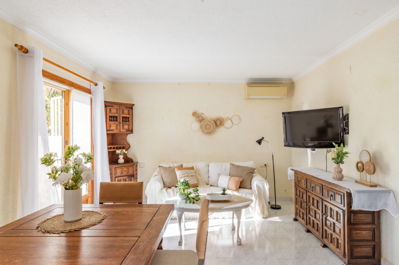 Salón-comedor luminoso con suelos cerámicos, mobiliario de madera rústica, sofá blanco y acceso directo al exterior a través de puertas acristaladas.