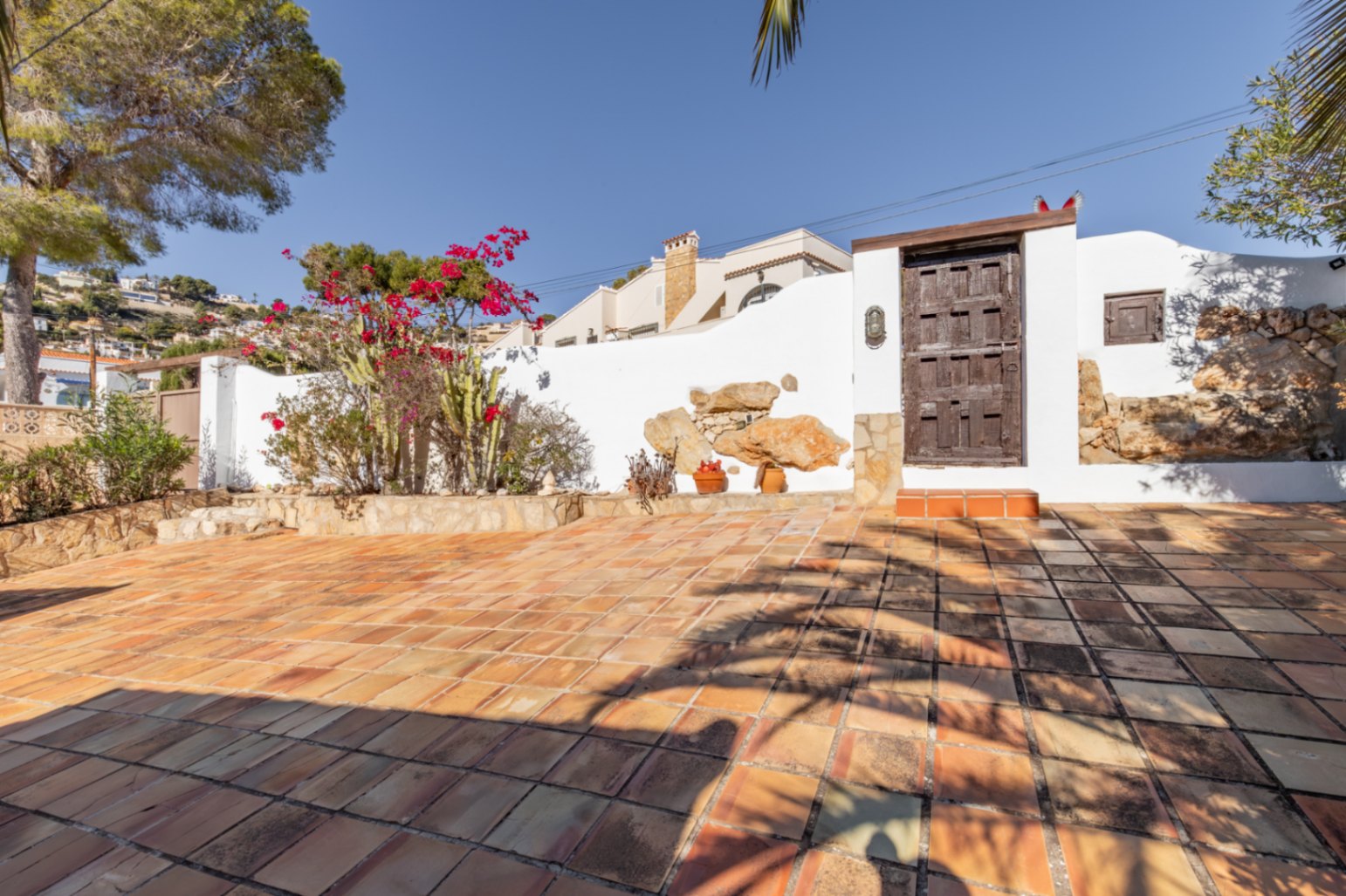 Terraza exterior con suelo de baldosas de barro, muros blancos con detalles de piedra natural y un portón rústico de madera rodeado de buganvillas y cactus.
