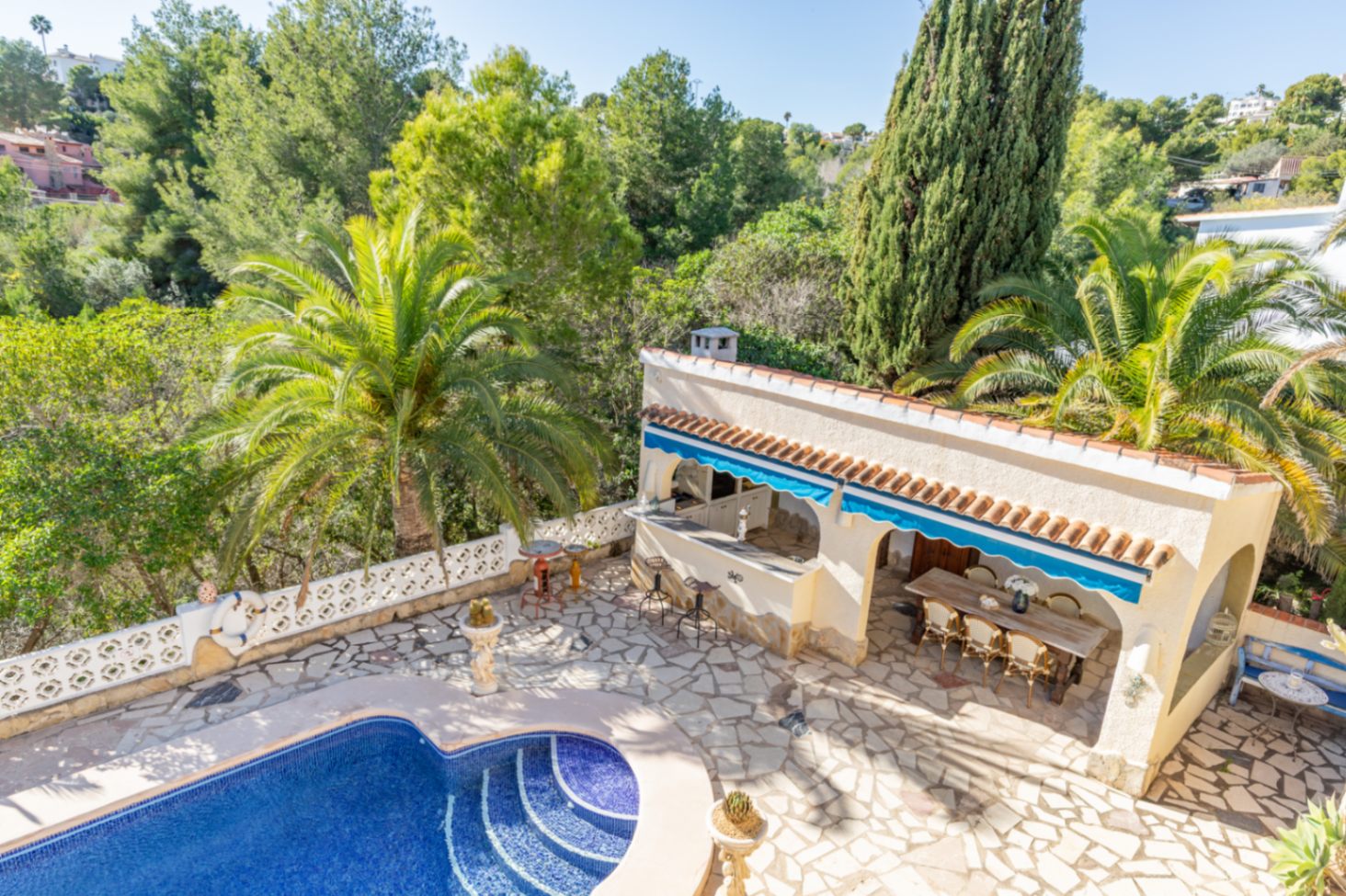 Zona exterior con piscina de mosaico azul, amplia terraza de piedra y cocina de verano techada, rodeada de palmeras y vegetación madura junto a la zona verde.