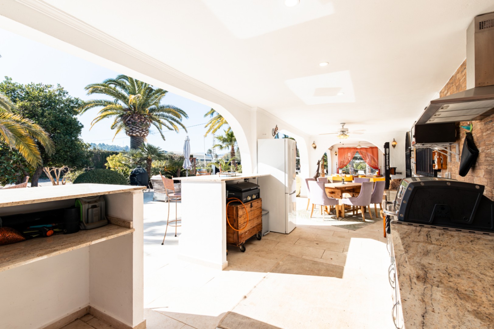 Amplia naya con cocina exterior equipada, encimeras de piedra y zona de comedor bajo arcos tradicionales con vistas al jardín mediterráneo.
