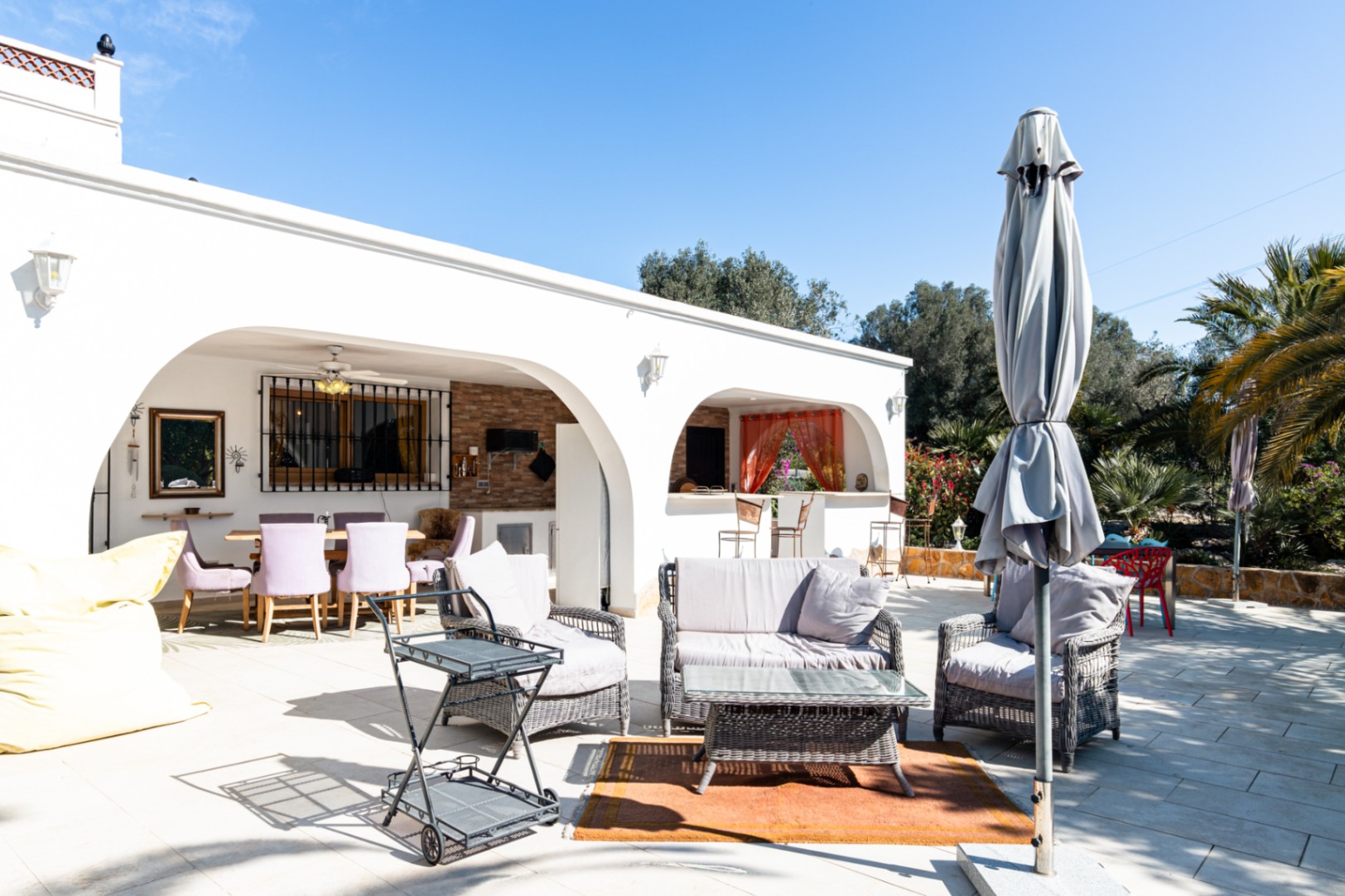 Amplia terraza exterior con mobiliario de mimbre, zona de estar y naya arqueada que alberga un comedor y cocina de verano bajo ventiladores de techo.