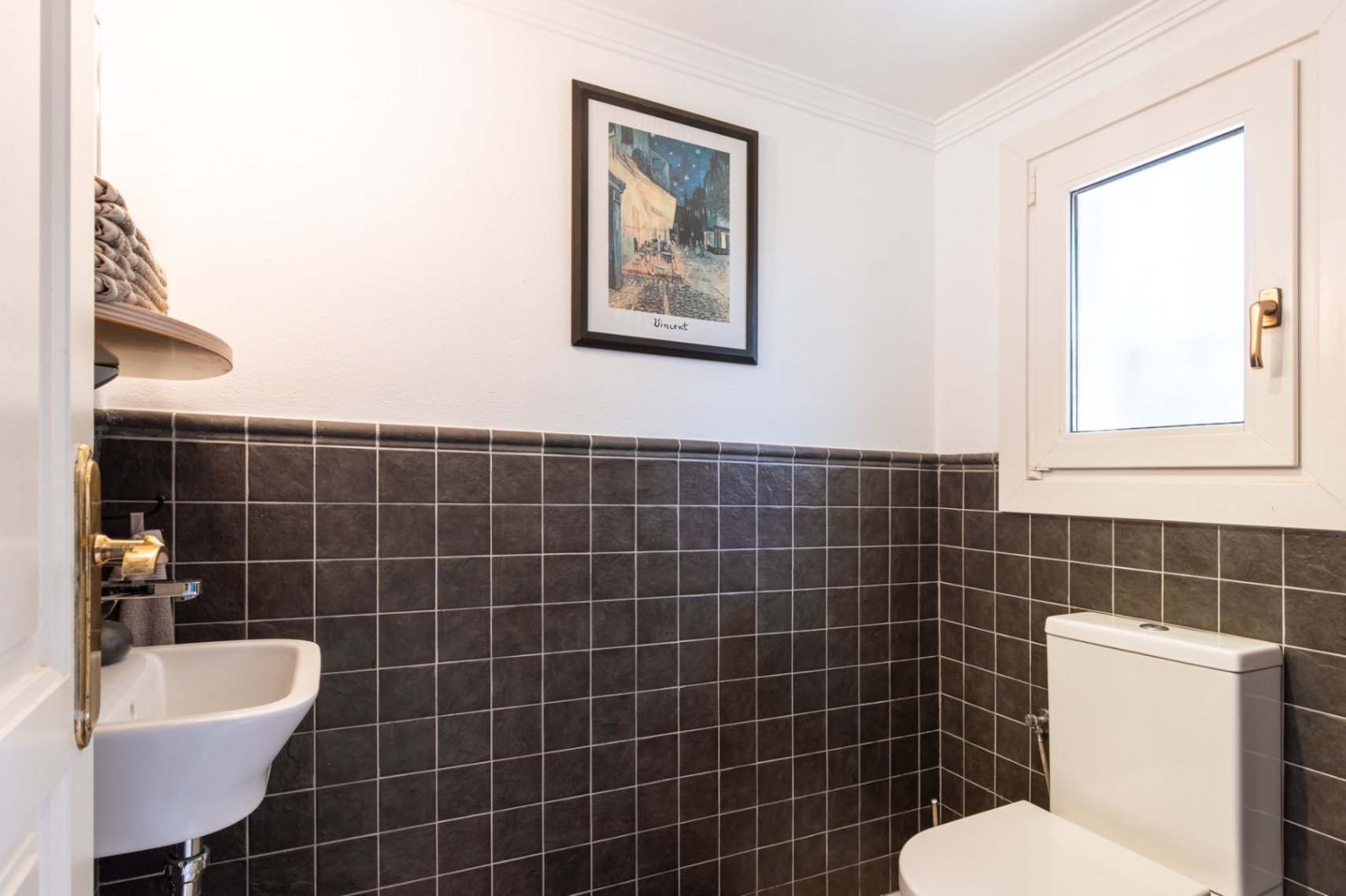 Aseo de invitados funcional con azulejos oscuros texturizados, lavabo compacto y ventana oscilobatiente para ventilación natural.