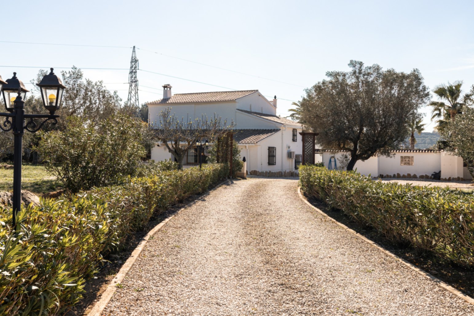 Camino de acceso de grava flanqueado por setos bajos que conduce a la fachada blanca de la finca, destacando su arquitectura tradicional y entorno mediterráneo.