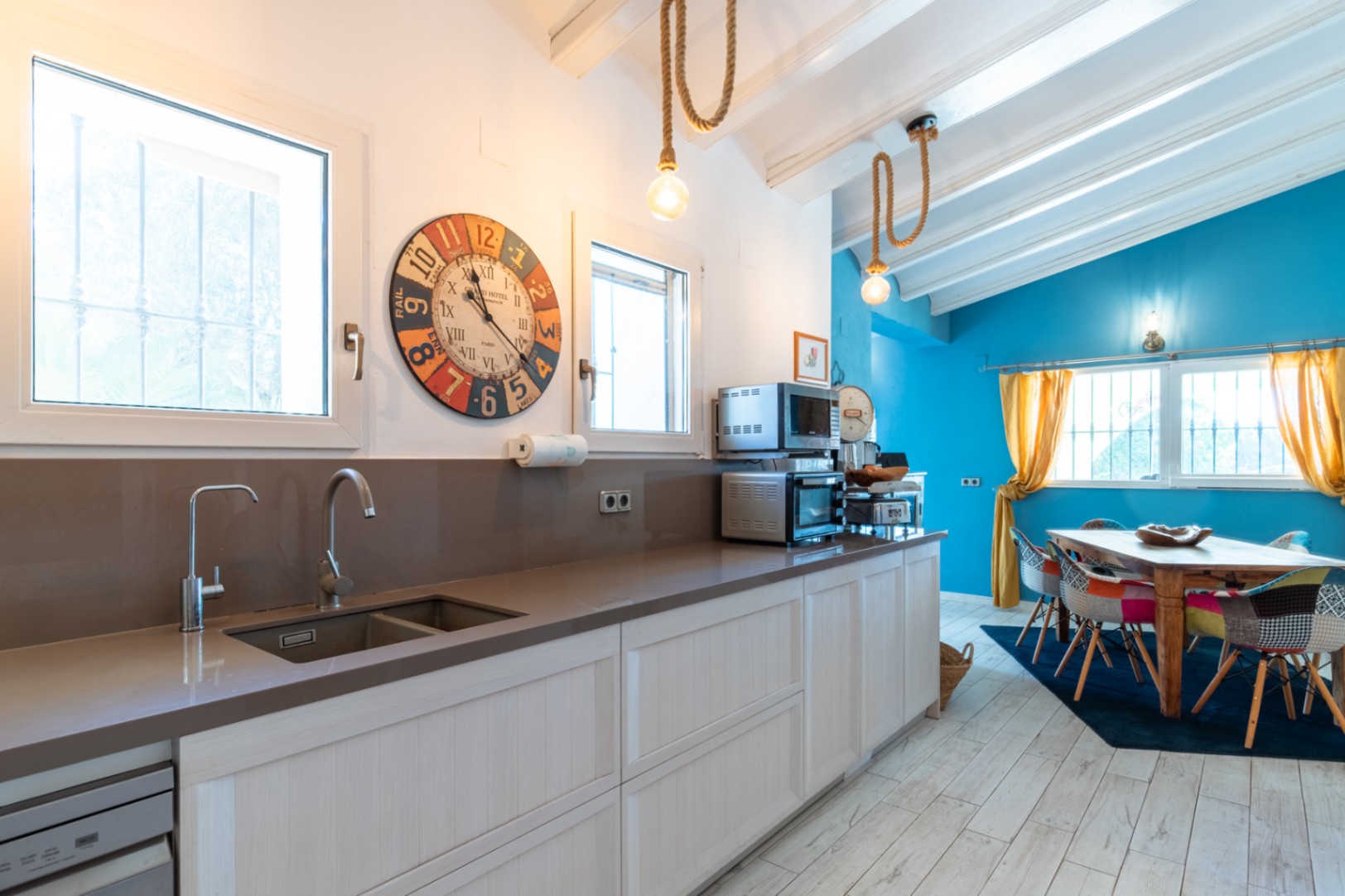 Cocina moderna con encimera de cuarzo y fregadero integrado, abierta a un comedor luminoso con paredes azules, vigas blancas y lámparas de cuerda de estilo rústico.