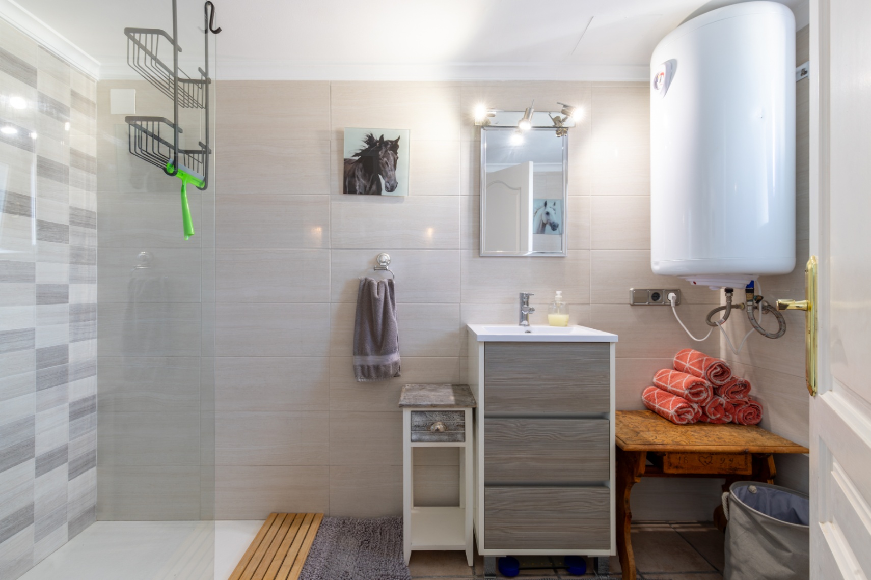 Cuarto de baño funcional con ducha a ras de suelo, mampara de cristal y azulejos texturizados. Dispone de lavabo con mueble gris, calentador eléctrico y detalles en madera.
