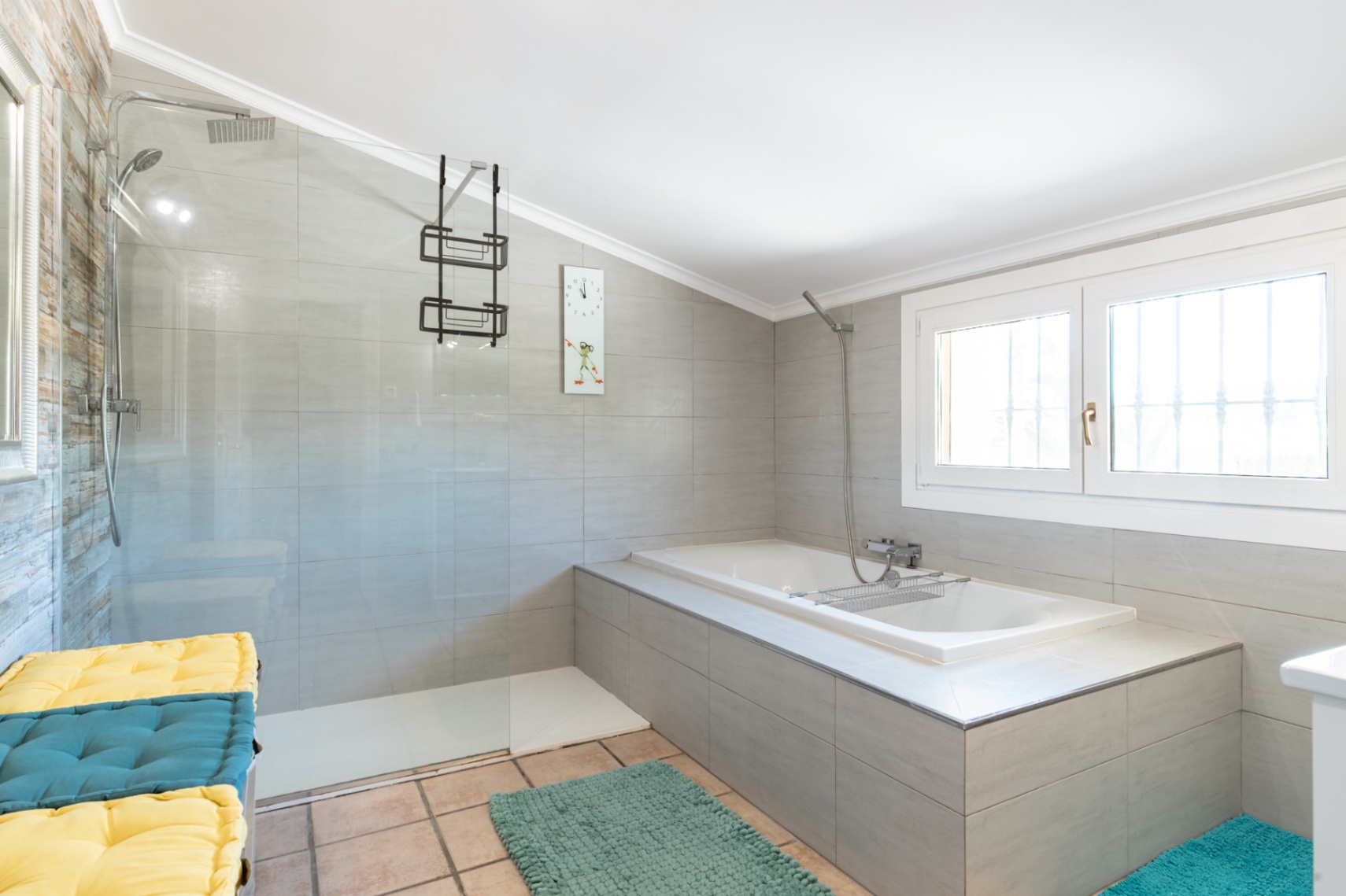 Cuarto de baño moderno con ducha a ras de suelo, mampara de cristal y bañera encastrada. Presenta revestimientos cerámicos en tonos grises y suelos de barro cocido.