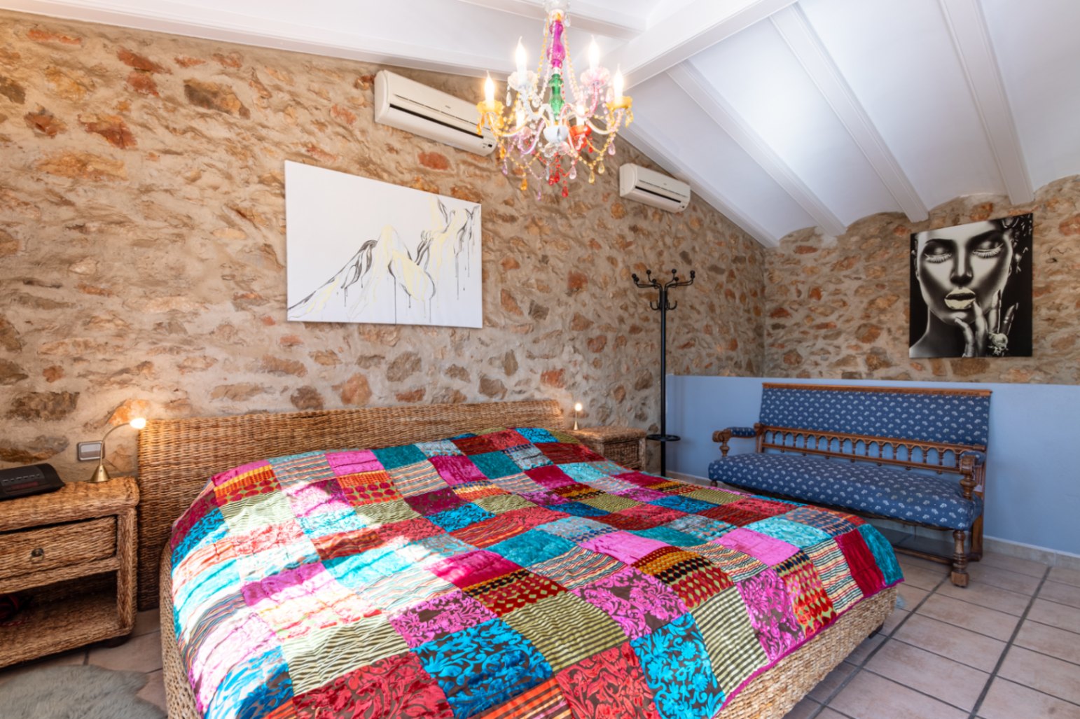 Dormitorio con paredes de piedra natural vista, techo de vigas blancas inclinadas y una cama de mimbre con colcha de patchwork bajo una lámpara de araña vibrante.