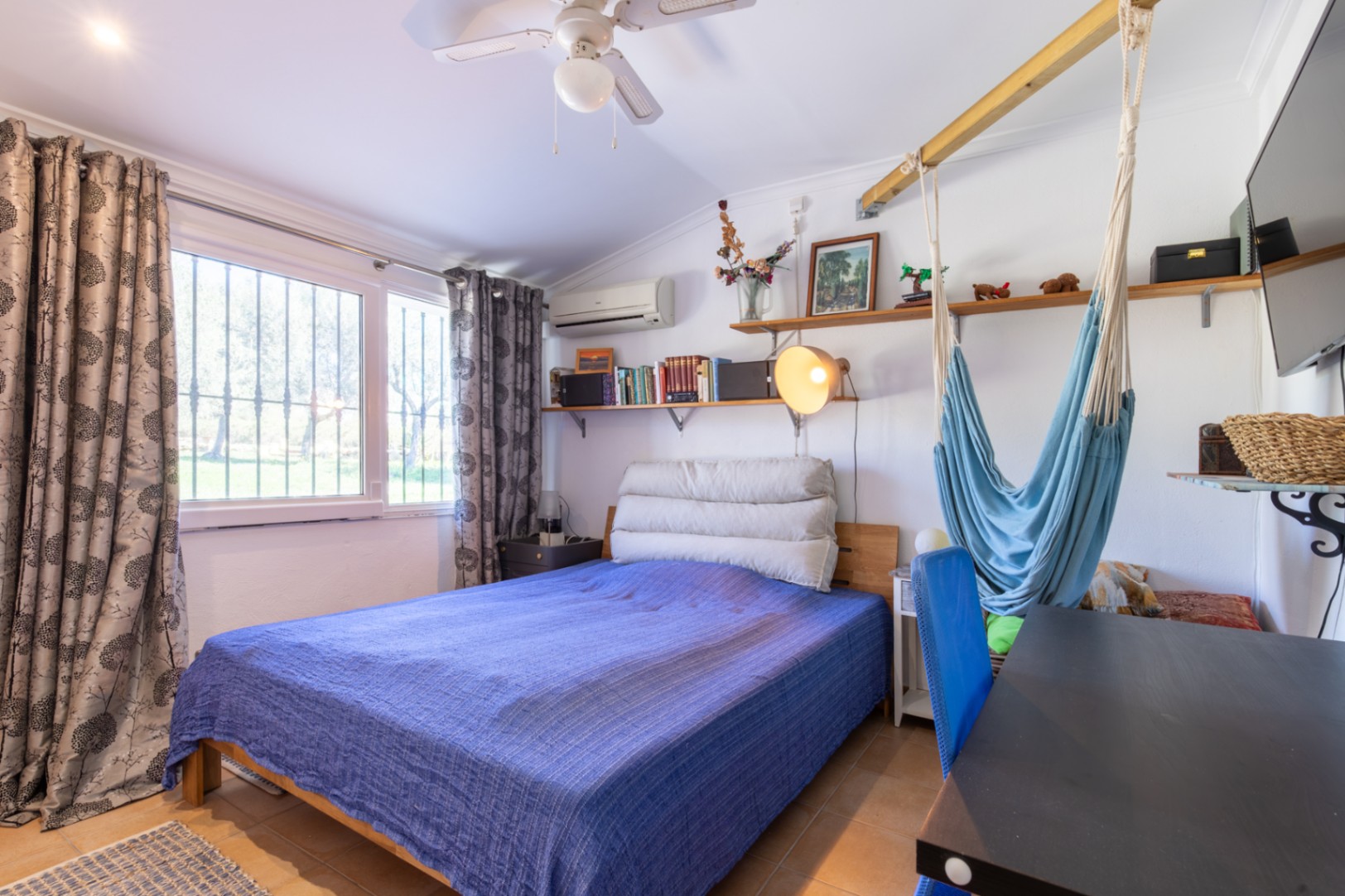 Dormitorio funcional con cama doble, zona de escritorio, estanterías de madera y una hamaca colgante. Cuenta con aire acondicionado, ventilador de techo y luz natural.