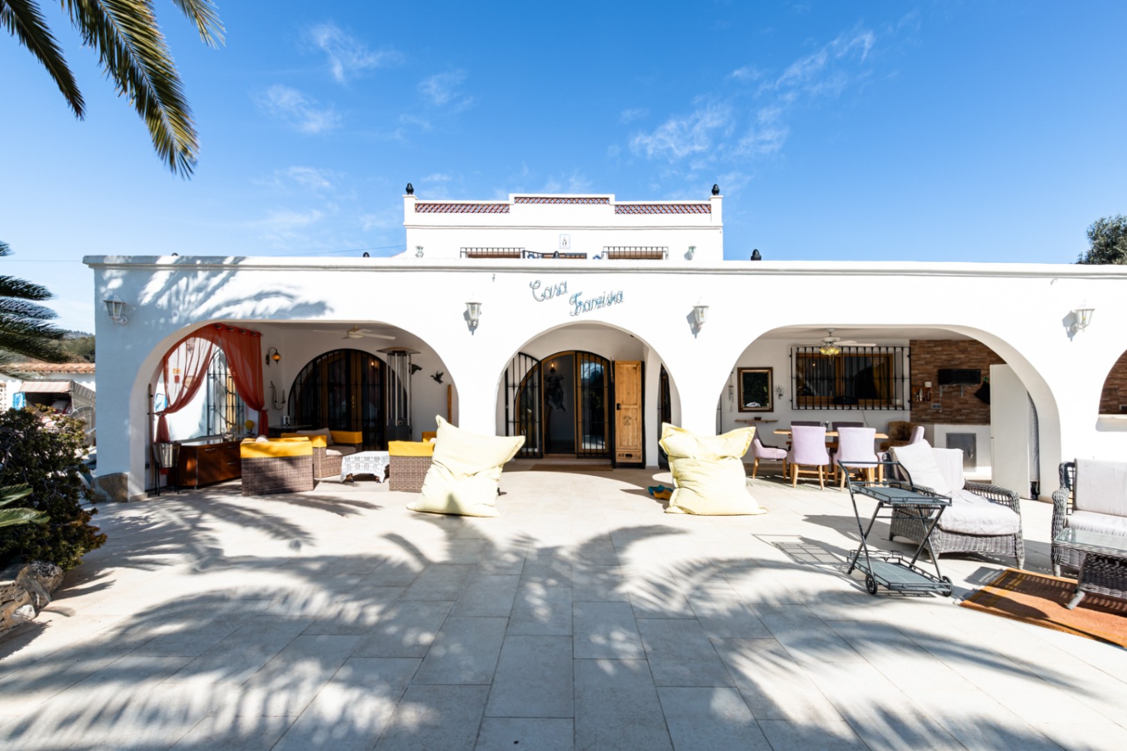 Fachada blanca de estilo mediterráneo con naya de arcos, amplia terraza pavimentada y zonas de estar exteriores bajo el rótulo "Casa Franziska".