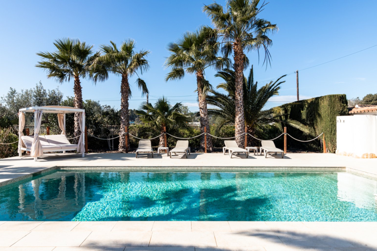 Piscina de 9,5 x 5 metros con zona de solárium pavimentada, equipada con tumbonas y una cama balinesa bajo la sombra de palmeras maduras.