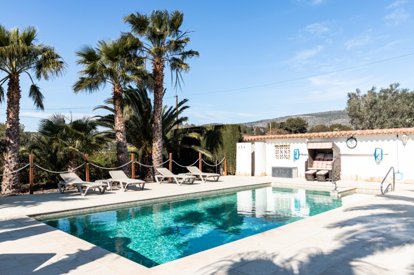 Piscina de 9,5m x 5m con terraza de piedra clara, zona de hamacas y palmeras maduras, integrada en un entorno privado con vistas despejadas hacia las montañas.