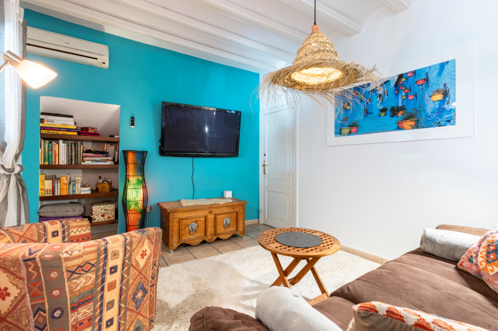 Salón acogedor con pared de acento turquesa, vigas de madera blancas y estantería de obra. Incluye aire acondicionado, mobiliario rústico y una lámpara artesanal de fibras naturales.