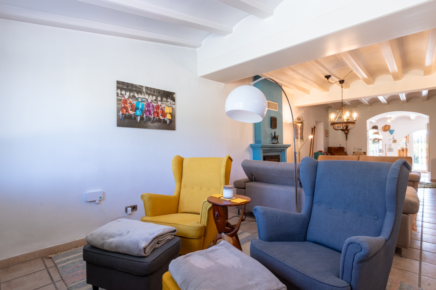 Salón acogedor con techos de vigas blancas, chimenea azul al fondo y sillones orejeros en tonos amarillo y azul junto a una lámpara de arco moderna.
