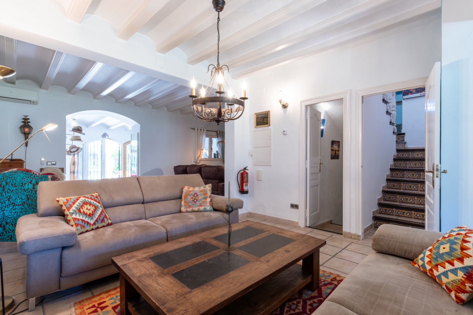 Salón con techos de vigas blancas, suelos cerámicos y escalera con azulejos tradicionales. Incluye una mesa de centro de madera maciza y lámpara de forja.