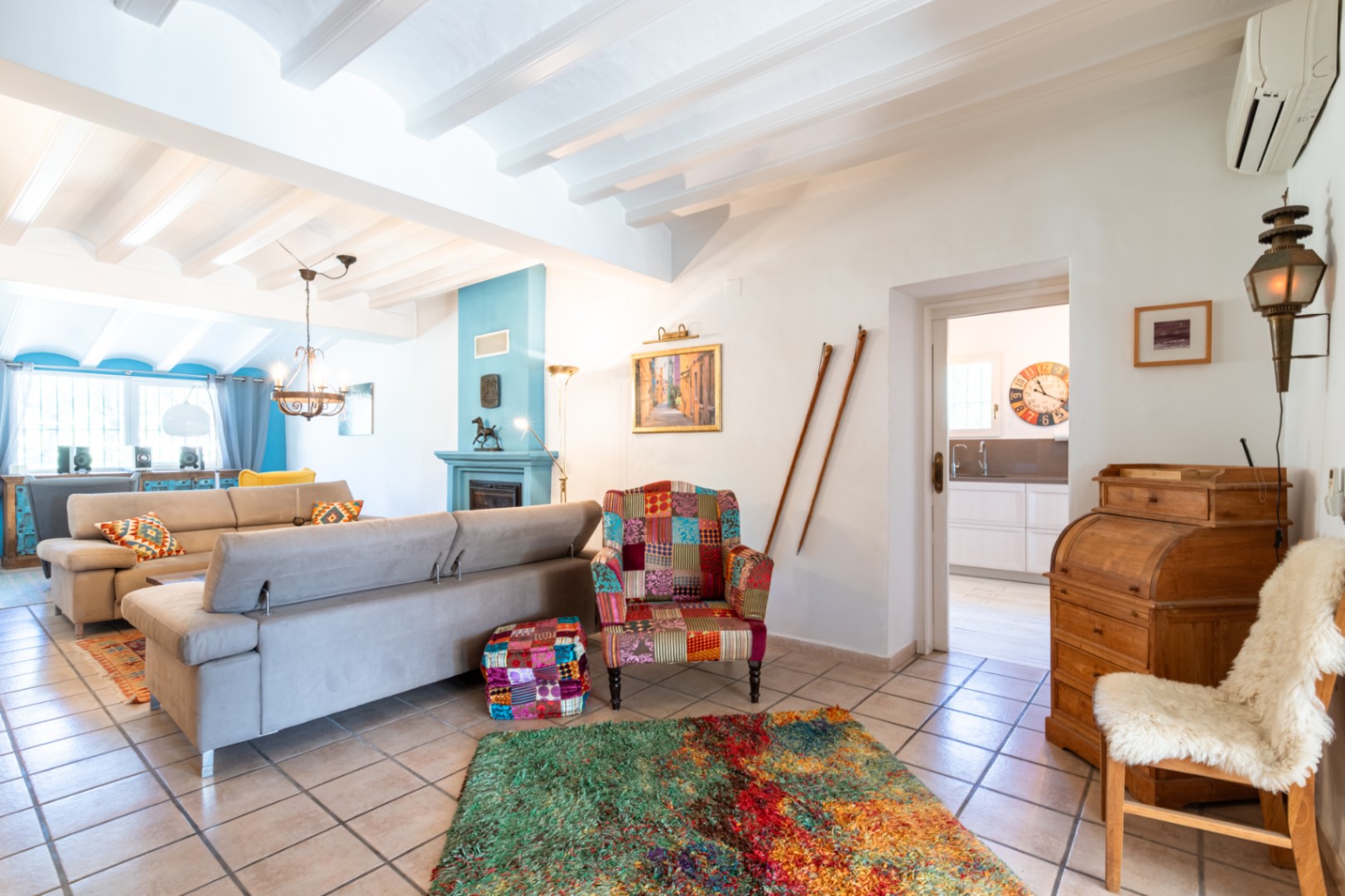 Salón luminoso con techos de vigas blancas, chimenea azul y mobiliario ecléctico. Incluye una amplia zona de estar con sofás modernos y acceso directo a la cocina.