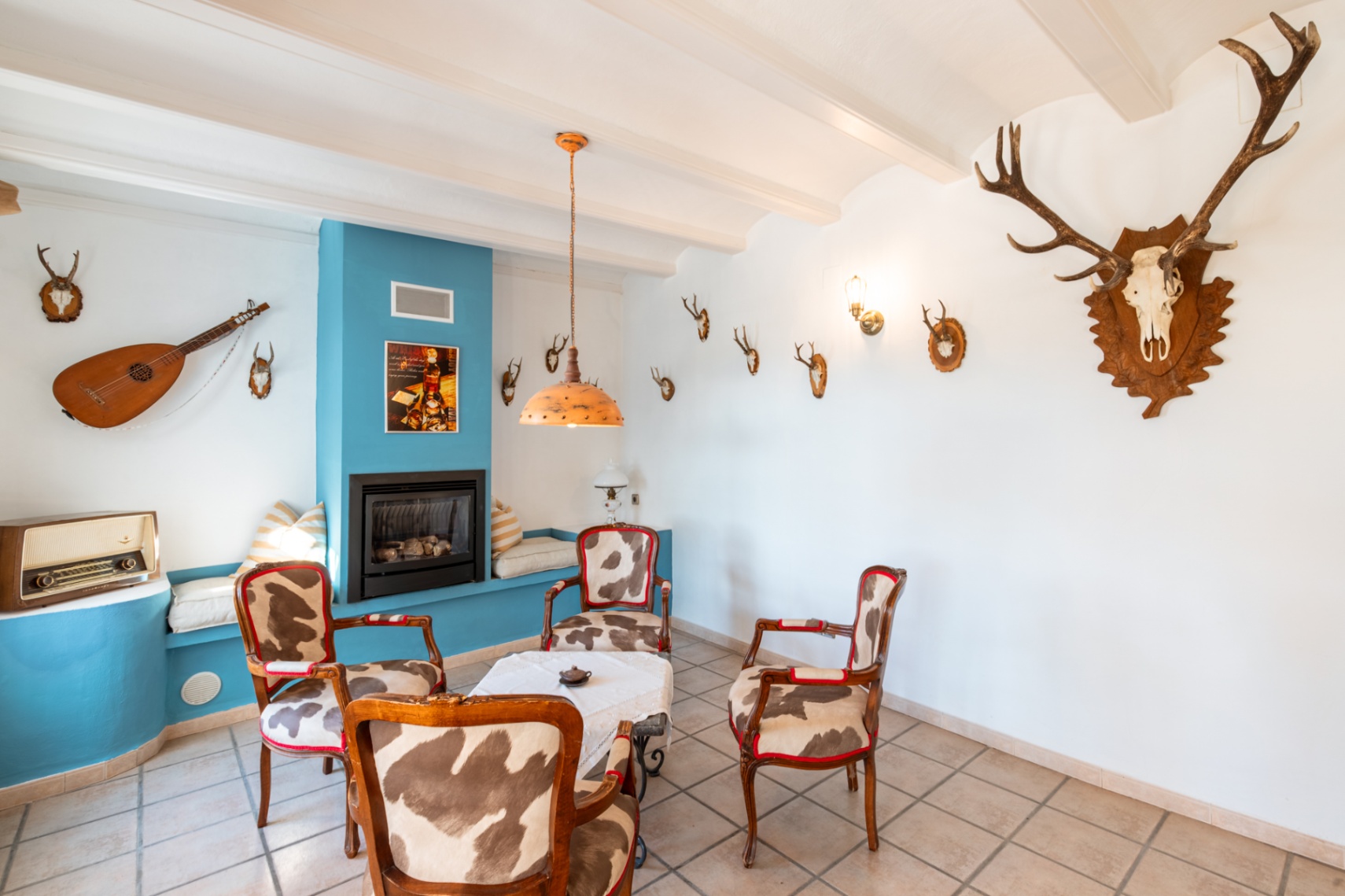Salón rústico con chimenea de gas integrada en pared azul, mobiliario tapizado en piel de vaca y techos con vigas blancas decorados con elementos cinegéticos.