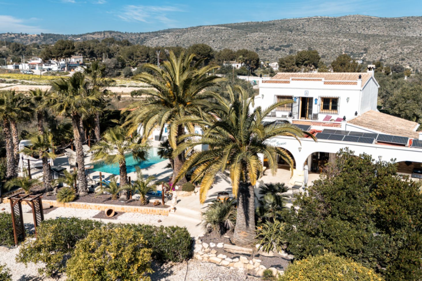 Vista aérea de la finca que destaca la instalación de paneles solares sobre la naya, la piscina privada rodeada de palmeras y el entorno natural de la parcela.
